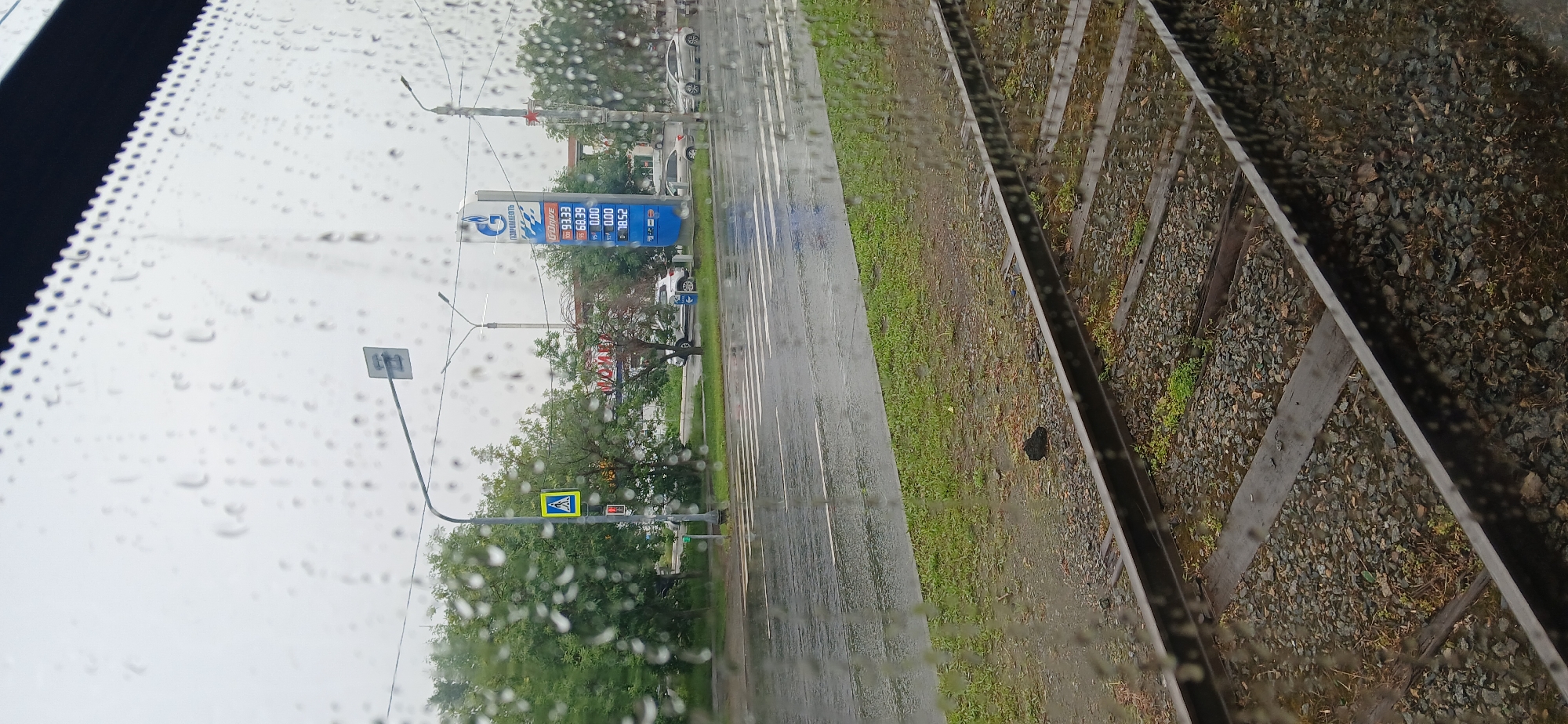 The image depicts a rainy day scene from inside a vehicle looking out a window covered in raindrops. In the foreground, train tracks run horizontally, with ballast and wooden sleepers visible. Beyond the tracks is a wet, grassy area, and then a wet asphalt road with white pedestrian crossing markings. Across the road, there are trees and a Gazprom gas station with a price board displaying fuel prices. Further back, buildings are visible, including one with the word "МОЙКА" (car wash) in large letters. Several cars are parked or driving on the road. A pedestrian crossing sign is also visible. The sky is overcast, suggesting cloudy weather. The scene appears to be a typical urban street with infrastructure and services. The location is identified as Chelyabinsk, Russia.