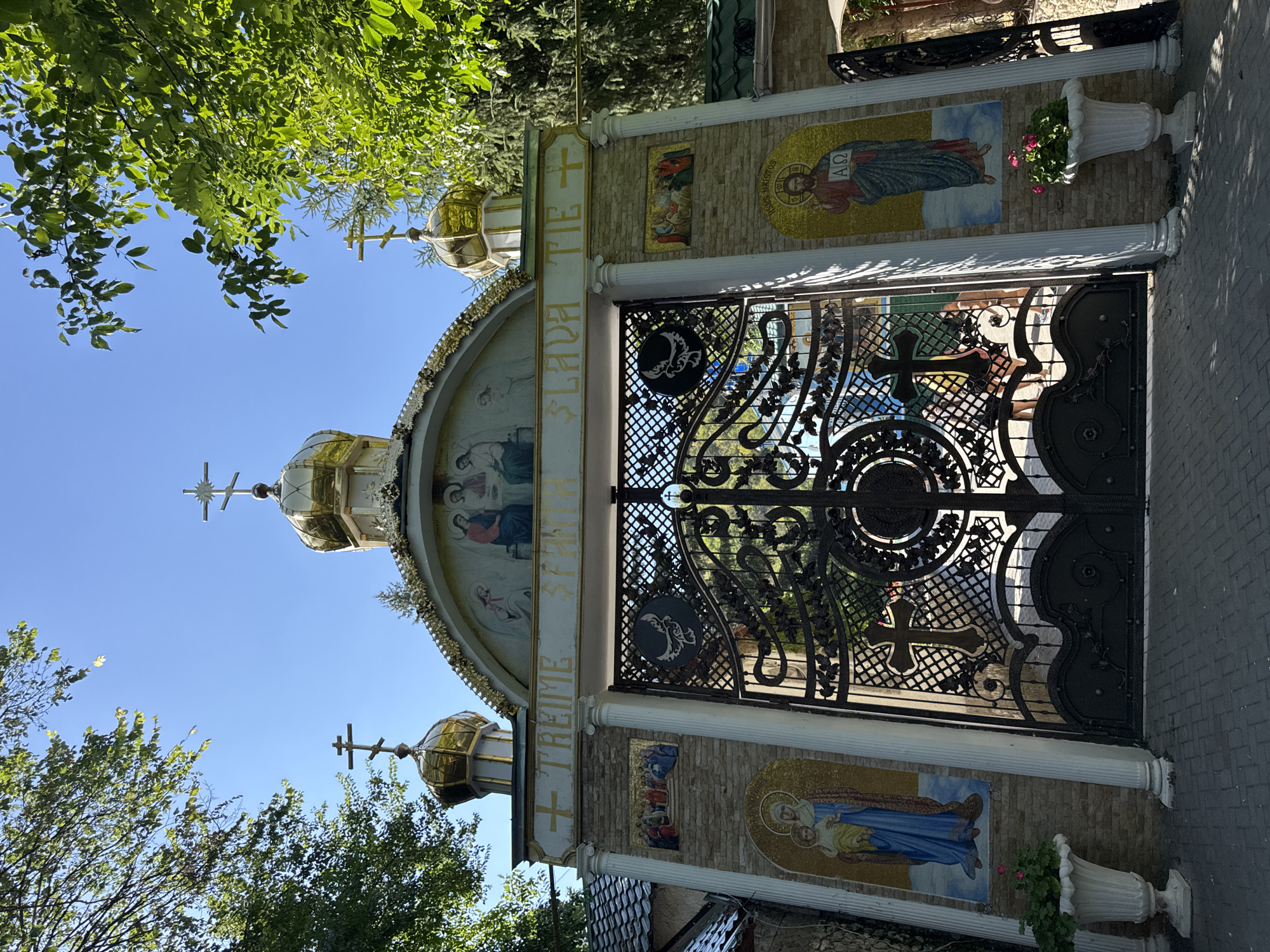 The image depicts the ornate entrance to a religious complex in Saharna, Moldova. A large, black wrought-iron gate, intricately decorated with floral patterns and a large cross, dominates the center of the frame. The gate is flanked by stone pillars adorned with mosaics. To the left, a mosaic depicts the Virgin Mary holding the Christ child, and to the right, a mosaic portrays Jesus Christ. Above the gate, an archway features a fresco of religious figures and a golden dome topped with a cross. Further behind the main gate, another section of the religious complex is visible, including more golden domes and crosses. The entrance is set within a courtyard paved with gray stones. Lush green trees frame the scene, and the sky above is a clear, bright blue. Two white planters with pink flowers are positioned at the base of the pillars on either side of the entrance.