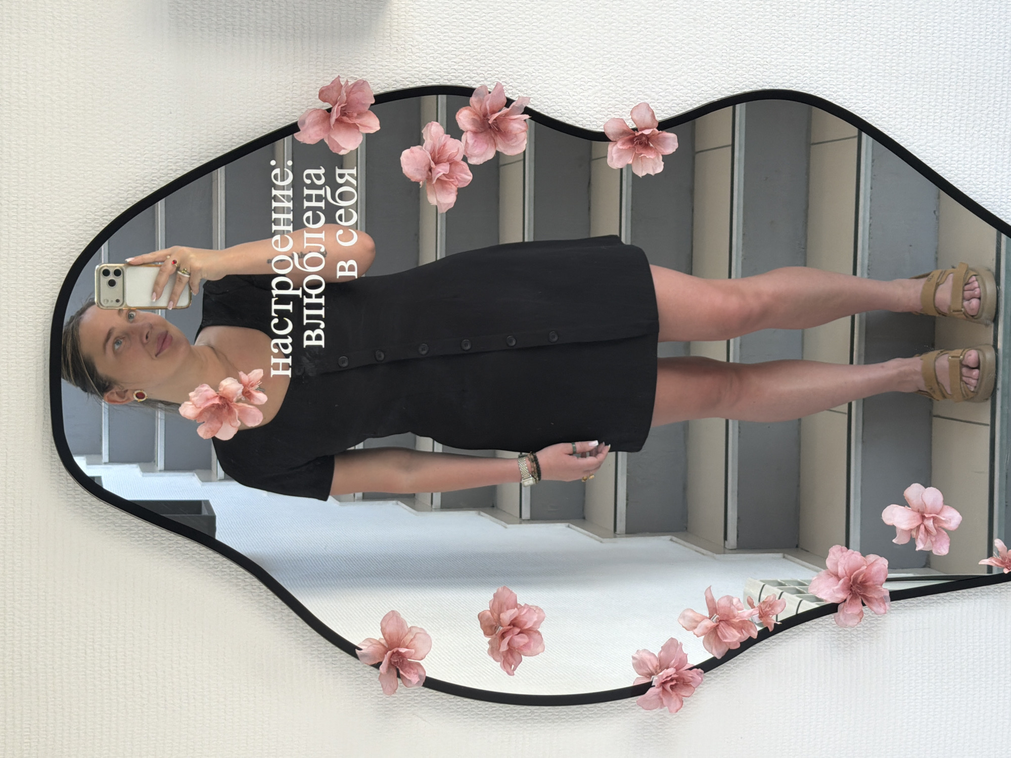 A woman is taking a selfie in a mirror. The mirror is oval-shaped with a black frame and has pink flowers attached around the frame. The reflection shows a staircase behind the woman. She is wearing a black dress and tan sandals. The text on the mirror reads "настроение: влюблена в себя" which translates to "mood: in love with myself." Based on prior knowledge, the location may be Chaikovsky, Russia.
