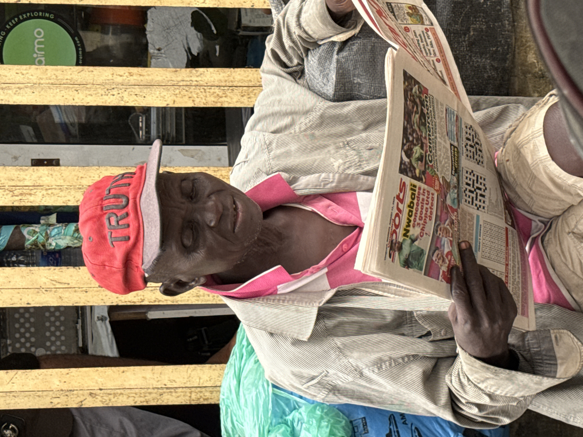An elderly Black man wearing a red "TRUTH" baseball cap, a pink and white striped shirt, and khaki shorts sits outdoors, holding and reading a newspaper titled "SPORTS". The man's head is tilted down towards the newspaper, and his eyes are closed or downcast. He is positioned in front of a yellow, slatted structure, likely part of a stall or building in a market or street setting. To his left, a large pile of bright green plastic bags is visible, with a blue plastic bag beneath them. The background includes more yellow slats and a glimpse of a shop or doorway. The overall setting suggests a busy urban environment. The newspaper has visible headlines and images of soccer players. The man's left arm is bent, holding the newspaper open. The photograph was taken in Lekki, Nigeria.