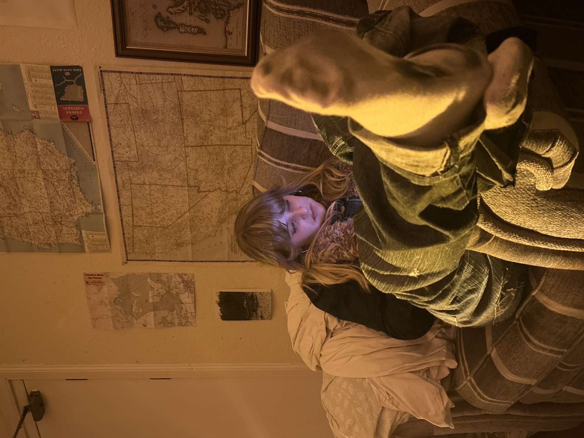 A candid, warm-toned photograph captures a person relaxing indoors, likely in Dundee, United Kingdom, during the evening or night. The subject, a person with blonde or light brown hair and bangs, lies on a striped couch or bed, their face illuminated by a bluish light, suggesting they are looking at a screen. Their feet, clad in light-colored socks, are casually propped up towards the viewer, slightly out of focus. They are dressed in dark trousers or jeans, a dark top, and a warm, possibly patterned, scarf or top around their neck.
The room's walls are adorned with several maps and framed artwork. Directly above the person's head hangs a framed picture, possibly an antique map. To the left, a vertical map is visible with "ESSO AUTO MAPA" and "ESPANA PORTUGAL" printed on its spine, alongside the number "140." A large, older-style map, potentially of a region in the "UNITED STATES" (with "CALIFORNIA" and "COLORADO" faintly discernible), covers a significant portion of the wall. Below this, a smaller, folded map appears to have text like "around & about" and "For Forest" printed on it. Another small, dark rectangular object, possibly a photo or artwork, is also on the wall. The overall ambiance is cozy and relaxed, with soft, warm lighting.