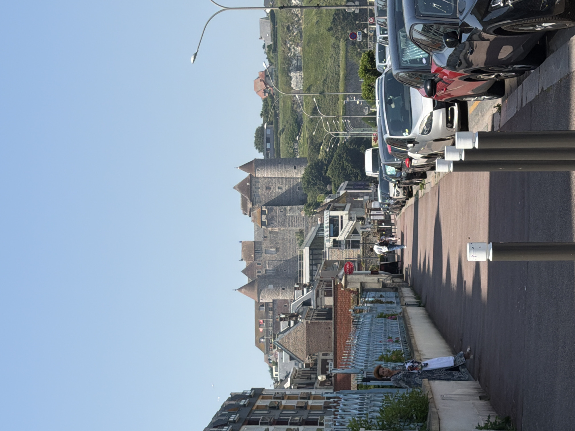 The image shows a street scene in Dieppe, France, with a view of the Chateau de Dieppe in the background. The Chateau, a large stone structure with towers and ramparts, is situated on a hill overlooking the town. In the foreground, a sidewalk runs alongside a street lined with parked cars. A woman with red hair, wearing a patterned dress, stands near a blue fence, holding a white bag. Several people are walking along the sidewalk and crossing the street. The sky is clear and blue, and the sun is shining, casting shadows on the ground. The buildings in the town are a mix of architectural styles, with some appearing older and historic, while others are more modern. The overall impression is of a sunny day in a historic coastal town.