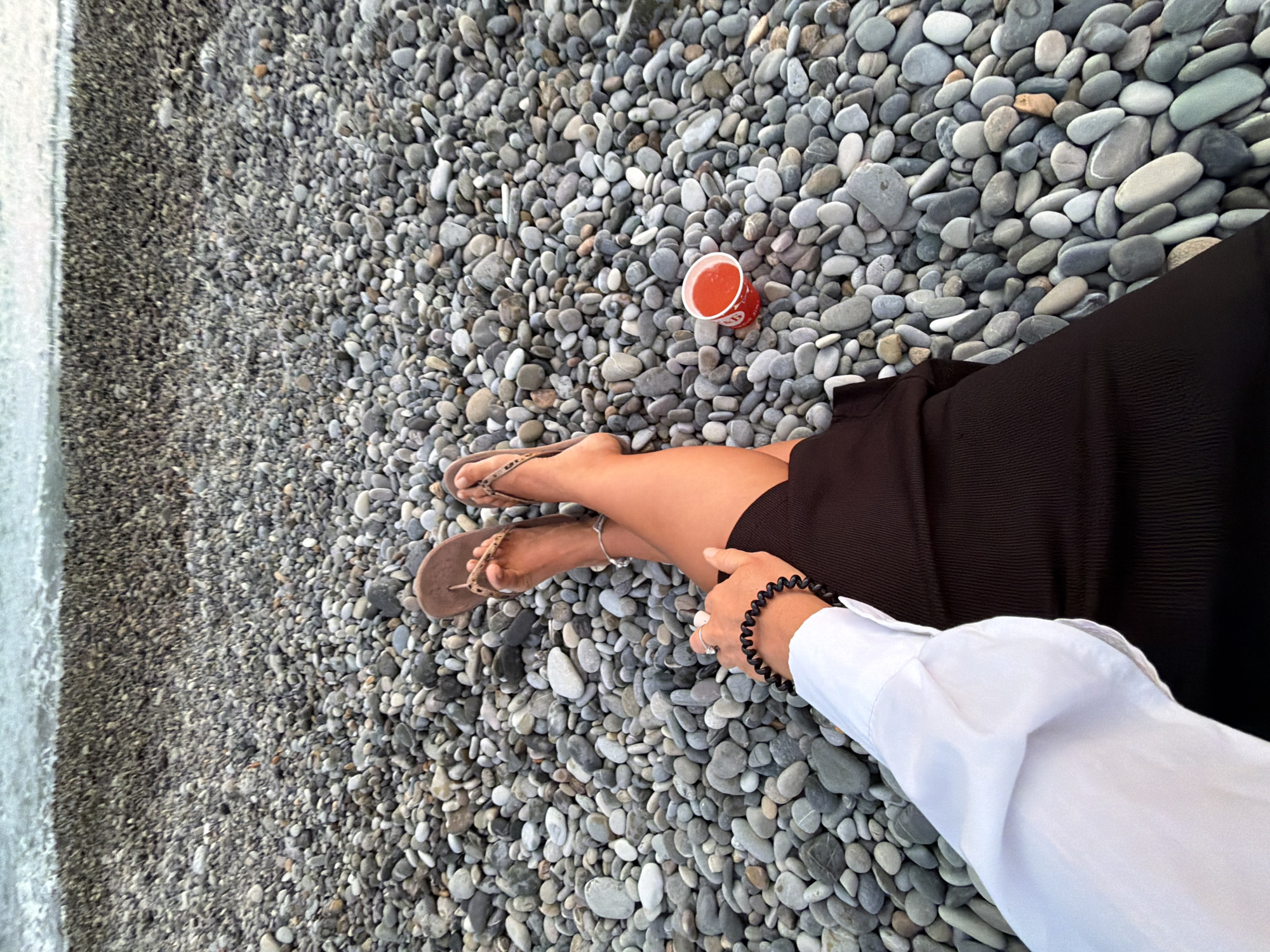 In an outdoor, beachfront location, a person is seated on a pebble beach with the ocean in the background. The person is wearing a white long-sleeved shirt, a black ribbed skirt, and brown flip-flops. They are also wearing a black beaded bracelet on their left wrist and a silver ring on their left index finger. A small red cup with a red liquid is placed on the pebbles to the person's right. The image is taken from a high-angle perspective, looking down at the person and the surrounding beach.