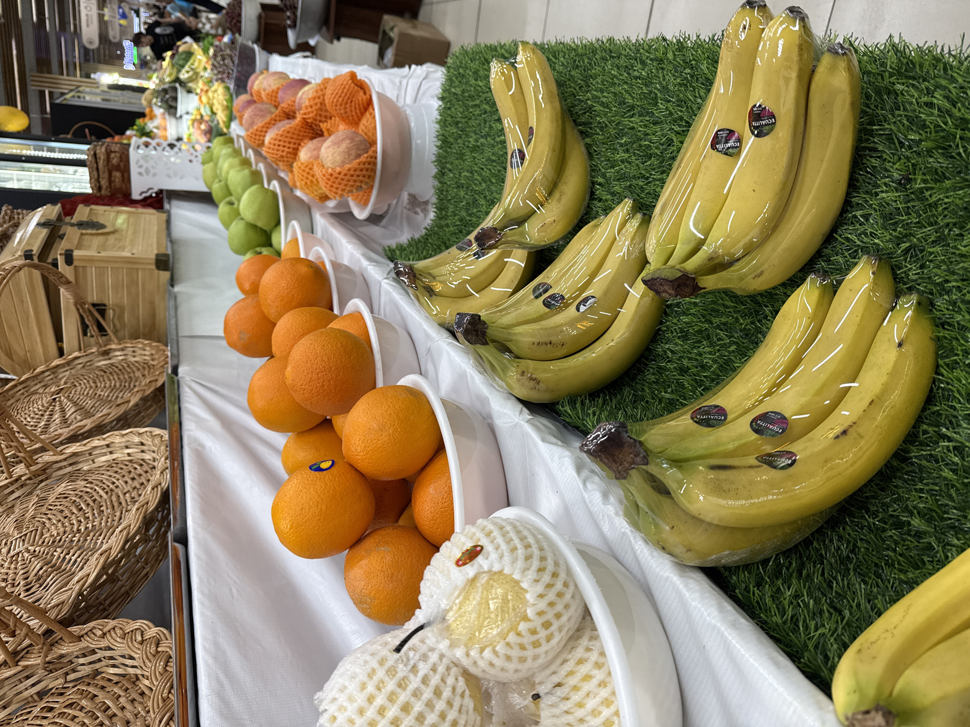 A display of fresh fruits is presented at a shop in Tashkent, Uzbekistan. The fruits are arranged in white bowls along a white counter surface. The display includes bananas wrapped in clear plastic, oranges, pears encased in protective netting, green pears, and peaches similarly protected by orange netting. The bananas rest on a strip of artificial turf. Wicker baskets are seen to the left of the counter, likely intended for customers. The fruit is situated indoors under artificial lighting.