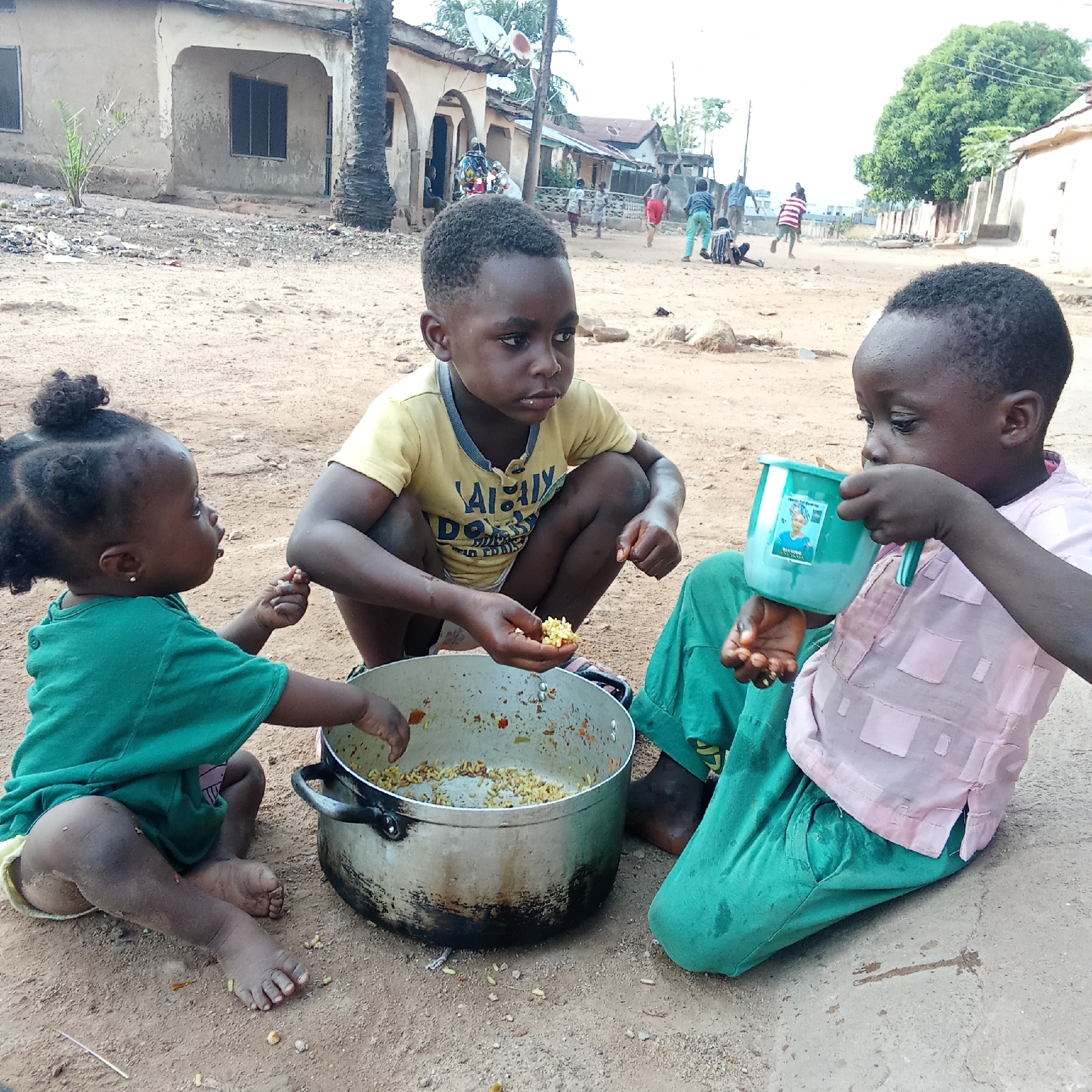 This outdoor scene in Akwanga, Nigeria, depicts three young children sharing a meal on a dirt ground. In the center, a boy wearing a yellow t-shirt with partially visible text "ALWAYS DO YOUR BEST" squats, holding a handful of a yellowish, grain-based meal, possibly rice, from a large, well-used metal pot. To his left, a baby or toddler with curly hair and a green top sits, reaching into the pot or holding a piece of food. To the right, another child in a pink patterned shirt and green pants drinks from a bright green plastic cup that has a sticker featuring a woman's image and some unreadable text.
The children appear focused on their meal, with neutral expressions. The ground is unpaved, dusty, and has scattered debris. In the background, several simple, somewhat weathered concrete buildings line an unpaved road. Palm trees and other sparse vegetation are visible. Other blurry figures, including adults and children, can be seen in the distance, suggesting a lively community setting. The bright lighting indicates it is daytime with clear weather. The overall environment suggests a humble, possibly rural or semi-urban, setting.