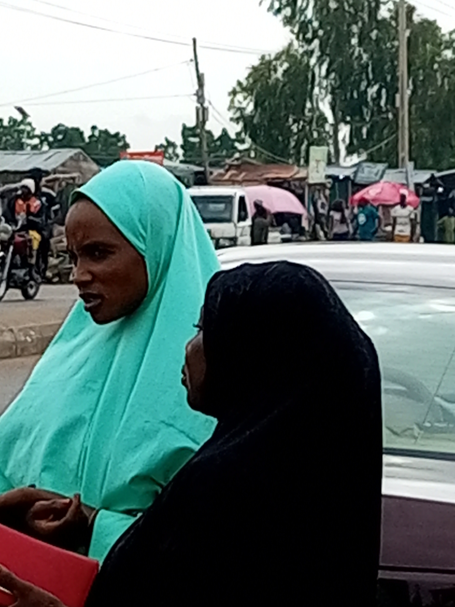 In Tempure, Nigeria, two individuals are captured in a close-up shot, partially obscured by the car they are near. On the left, a person wearing a teal hijab is facing left, with their mouth slightly open as if speaking. Their right hand is visible, holding a red object. To the right of this person, another individual is wearing a black hijab, and their face is partially visible from the side, looking to the left. The background reveals a busy street scene with various stalls, people, a white van, and trees. A motorcycle is also visible to the far left. The overall lighting suggests an outdoor setting, possibly a market.