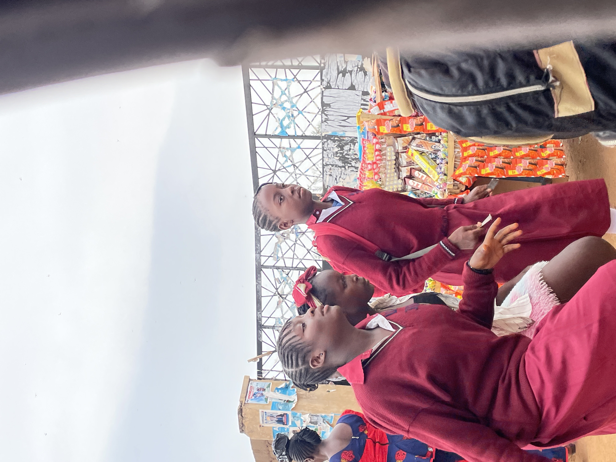 Three young schoolgirls are standing in front of a market stall in Jos, Nigeria. The girls are wearing matching maroon school uniforms. They have braided hair and are looking upwards. In the background, there is a market stall with various goods on display. The sky is overcast.