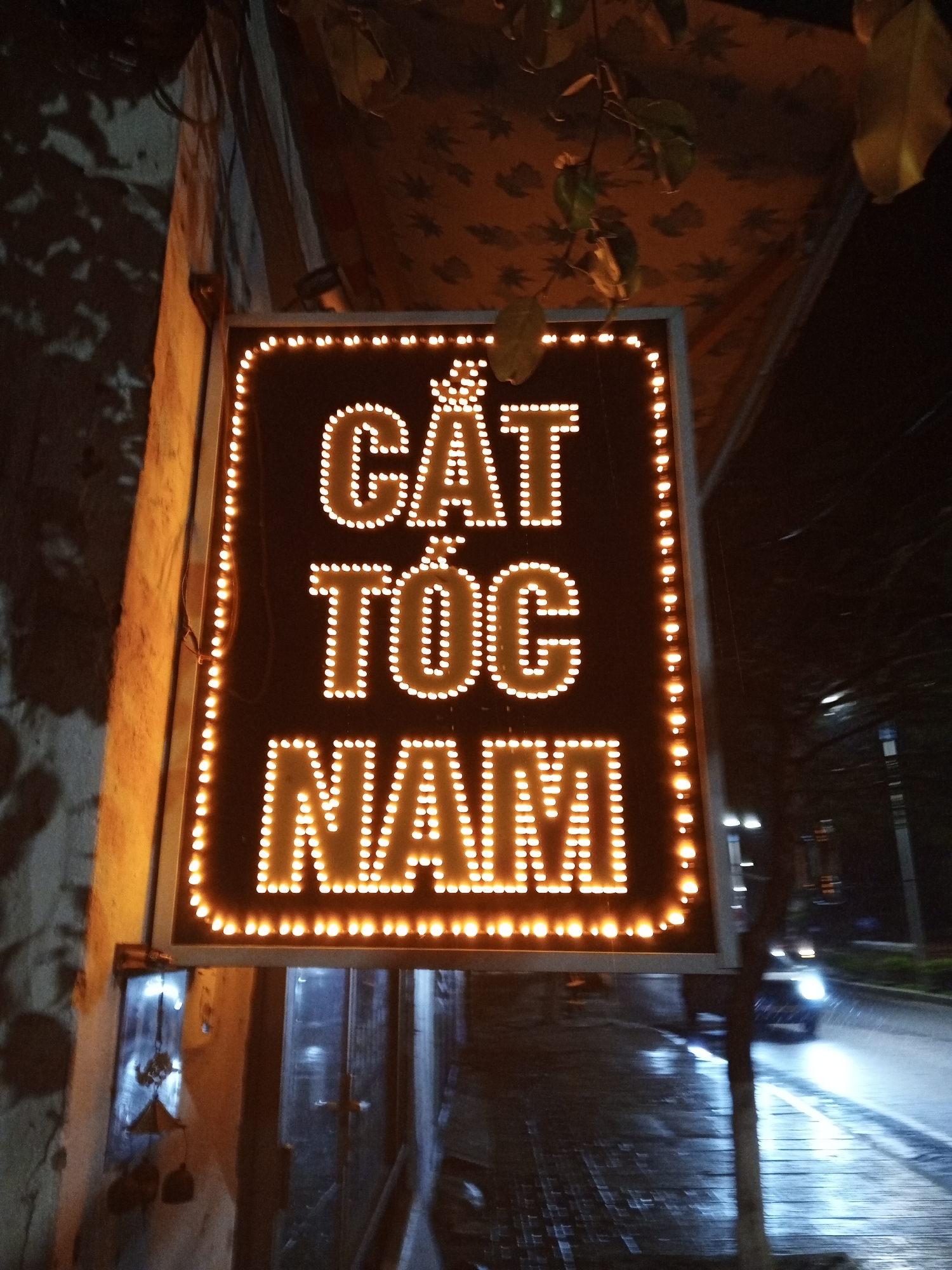 At night, a neon sign advertising "Cắt Tóc Nam" (men's haircut) is prominently displayed in Bac Nha Trang Ward, Vietnam. The sign, illuminated with bright lights, hangs near a building and a street. A car passes by on the wet road.