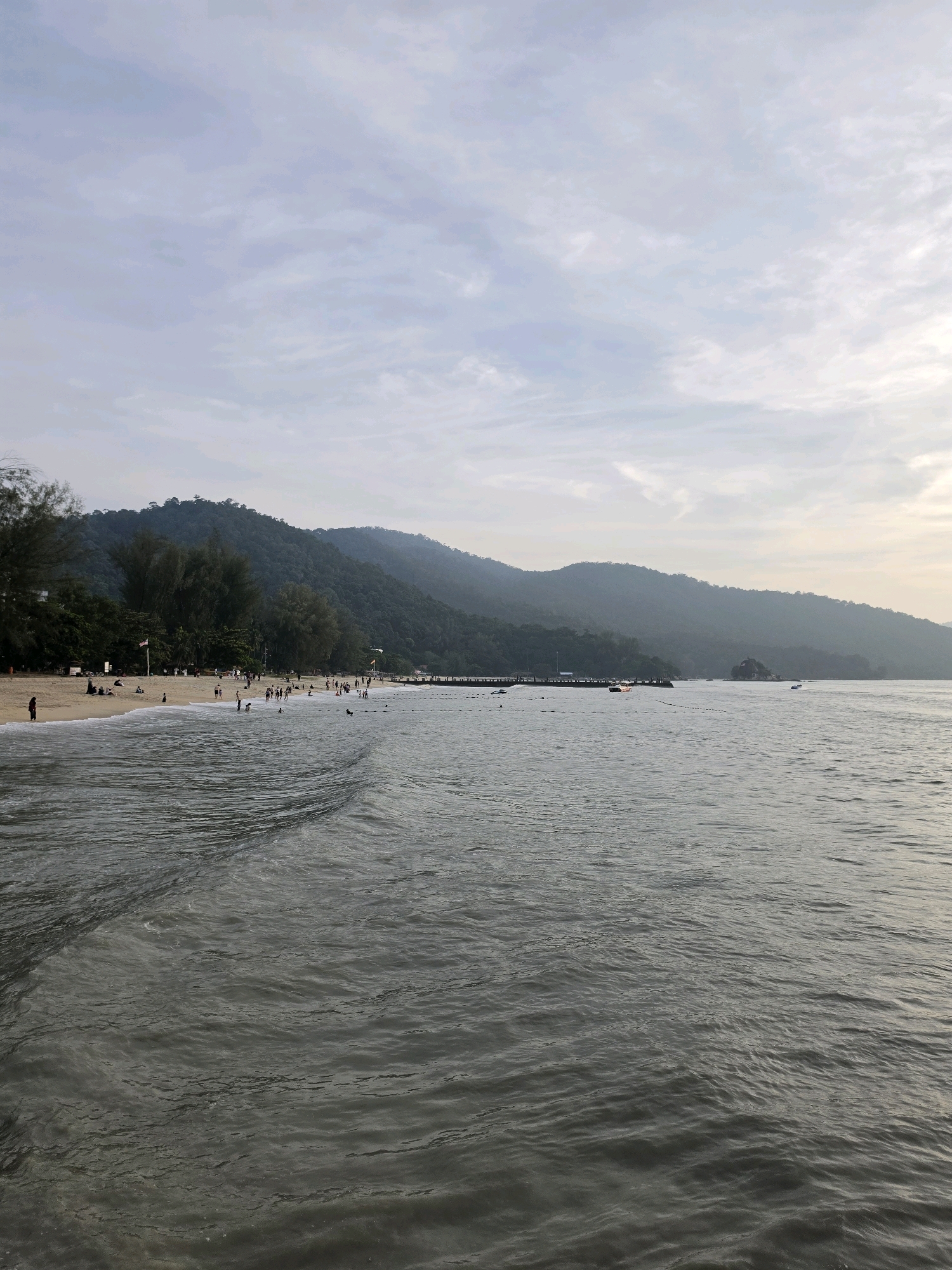 The image depicts a scenic view of a beach in George Town, Malaysia, likely Batu Ferringhi beach, known for its family-friendly atmosphere and watersports. The foreground is dominated by the calm, murky waters of the sea, with gentle waves lapping the shore. The middle ground features a sandy beach, populated by a moderate number of people engaged in various activities. Some individuals are seen walking along the shoreline, while others are enjoying the water, swimming or wading. A few are sunbathing or relaxing on the sand. In the background, a line of lush green trees and dense foliage forms a backdrop to the beach, leading up to the rolling, mist-covered hills that characterize the island's interior. The sky above is a soft, hazy blue, with wispy clouds indicating a peaceful, possibly late afternoon or early morning setting. A pier or jetty extends into the water on the right side of the image, and a few small boats are visible further out at sea, suggesting recreational boating activities. The overall impression is one of a tranquil coastal scene, characteristic of a popular tourist destination.
