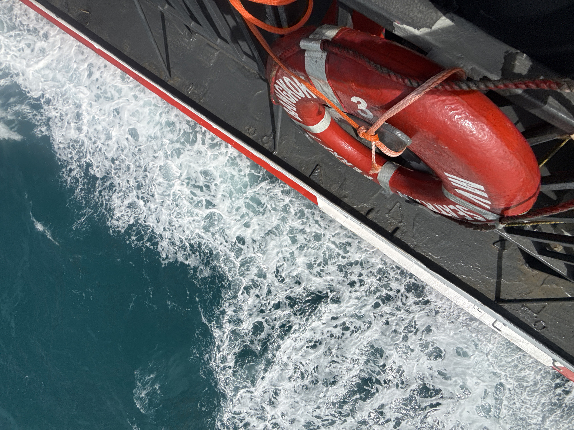 A high-angle, close-up shot captures a bright red life preserver on the deck of a vessel. The life preserver, with white lettering that appears to read "BANGKOK" and "NAV", is secured by orange and white ropes. The vessel's deck is dark gray and appears wet, with metal railings visible. To the left of the vessel, the turquoise water of the ocean churns with white foam, indicating movement. The border between the vessel and the water is marked by a white stripe with a thin red line. The scene suggests the vessel is in motion through the ocean. The location is the sea off the coast of Bangkok, Thailand.