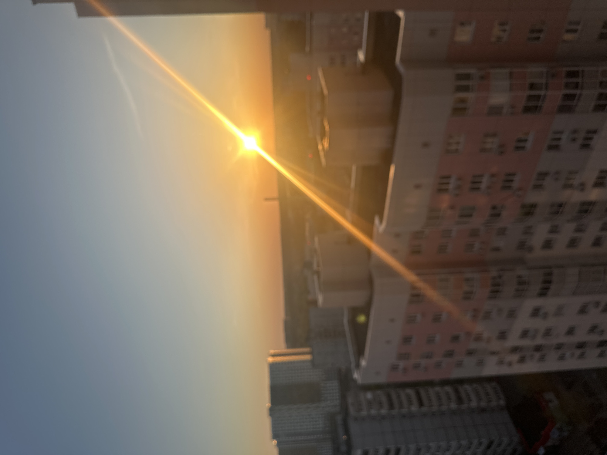 The image shows a sunset view of Moscow, Russia. The sun is low on the horizon, casting a warm glow across the sky and buildings. There are several high-rise buildings visible, some with pink and white facades and rows of windows. A portion of the sky is clear and blue.