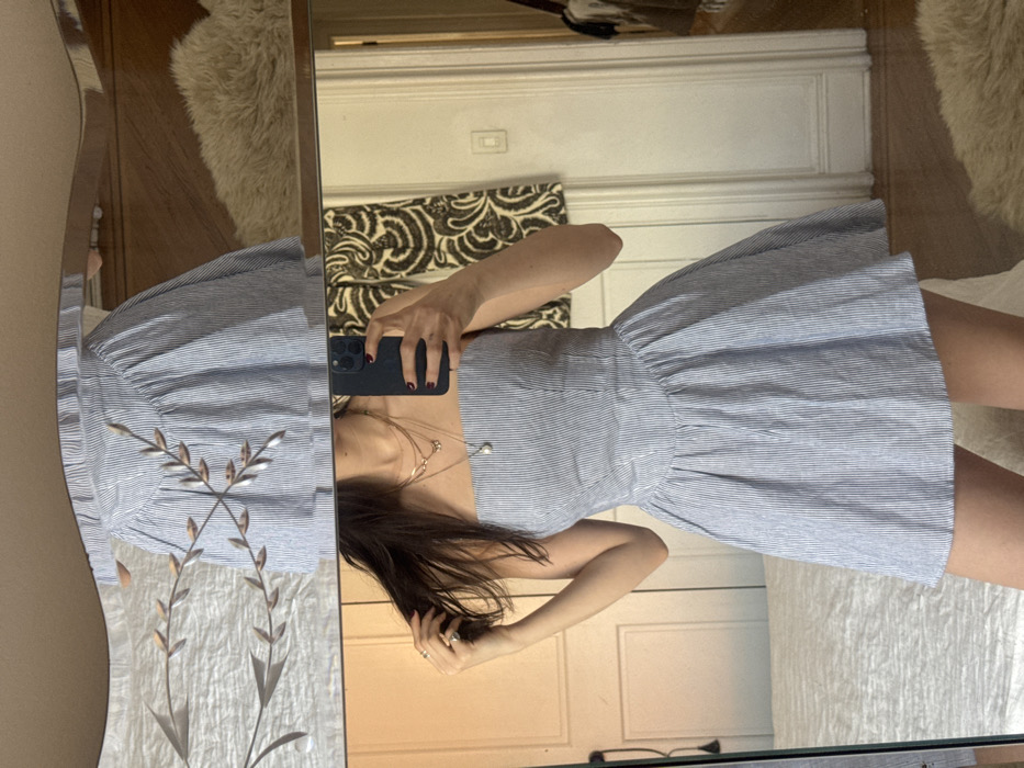 A person, likely female, with long dark brown hair, is depicted from mid-torso to mid-thigh, taking a mirror selfie in an indoor setting. The individual holds a dark-colored smartphone with three rear camera lenses in their right hand, obscuring their face. Their left hand is positioned near their head, gently touching a section of their hair.
The person is wearing a strapless dress with a fitted bodice and a pleated, flared skirt, featuring light blue and white vertical stripes. Multiple layered, delicate necklaces are visible on the neck, including one with a small pearl pendant and another with a small butterfly charm. Rings are worn on the fingers of the left hand.
The scene is reflected in a large, rectangular mirror. The top edge of the mirror is decoratively wavy and adorned with a metallic leaf-and-branch motif. The reflected environment suggests a bedroom or dressing area. To the left, a portion of a white or light-colored bed with a textured duvet is visible, alongside a white paneled door with a dark doorknob. To the right, a light-colored wooden floor, a cream-colored shaggy rug, another white paneled door, a white wall-mounted light switch, and a dark-patterned curtain or tapestry are present. Beyond the open door on the right, an interior space containing what appears to be hanging garments is partially visible.
The specific location within New York, United States of America, cannot be determined from the visual information provided.