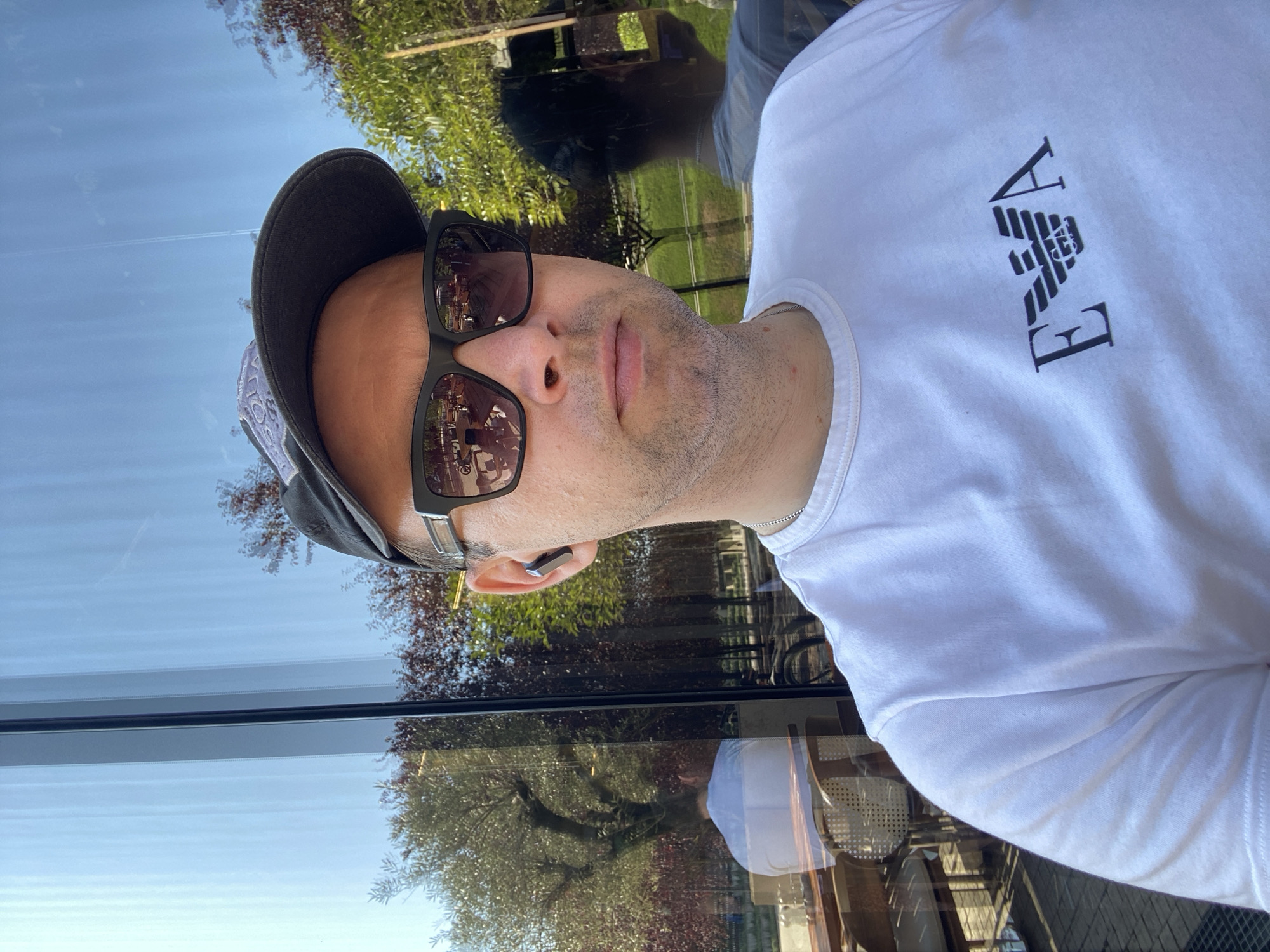 The image shows a close-up selfie of a man wearing a white t-shirt with the Emporio Armani logo, a dark baseball cap, and sunglasses. He has an earbud in his left ear. The man is sitting in front of a large window that reflects the sky, trees, and buildings, suggesting an outdoor or indoor seating area with a view. The reflection also shows another person sitting at a table in the background, as well as a table and chairs outside. The overall scene appears to be a cafe or restaurant. The man's attire and the environment suggest a casual setting during the daytime. There is no specific indicator that this location is in the city of Krasnodar, Russia.