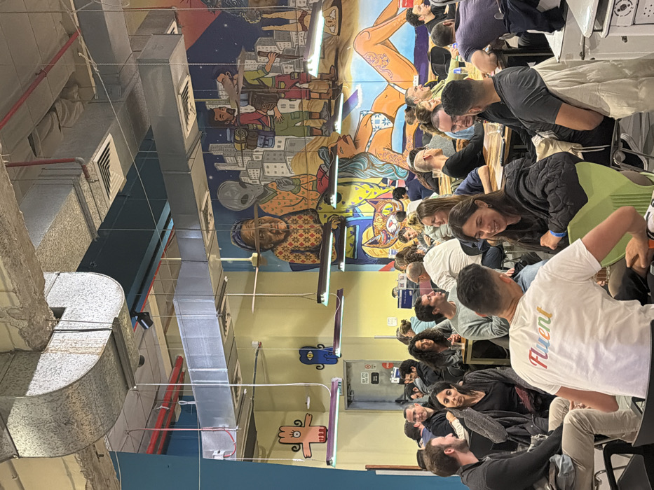 A room filled with people seated at tables, engaged in conversation and activity. The walls are adorned with colorful murals featuring diverse characters and scenes. Overhead, exposed ductwork and lighting fixtures run along the ceiling. A small cartoon monster is painted on the wall. The setting appears to be a casual, collaborative workspace.
