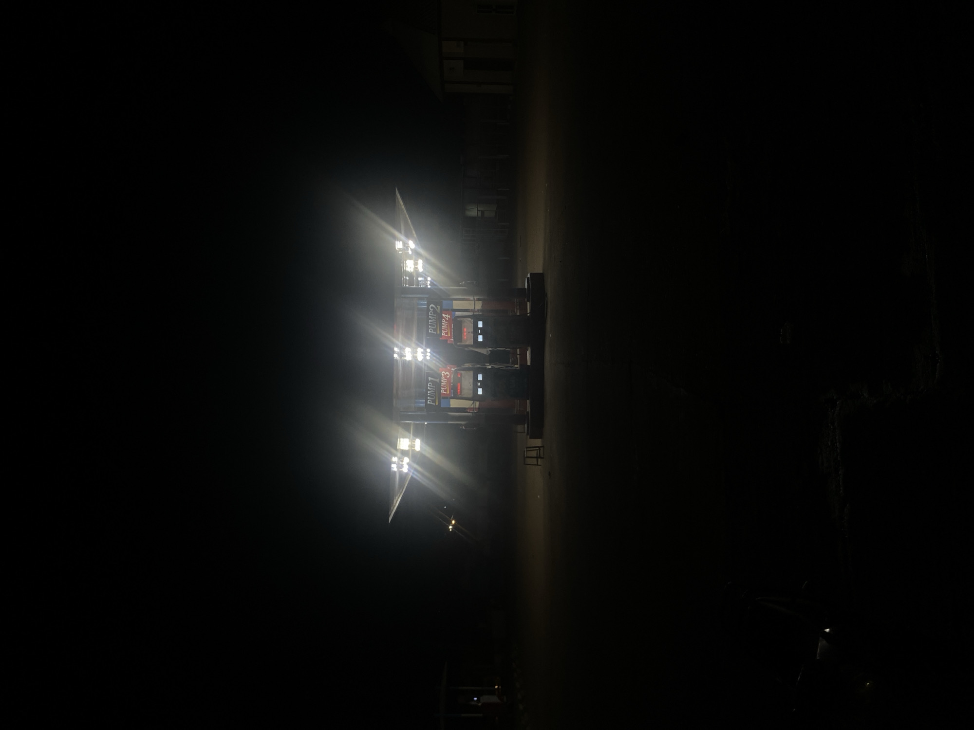 At night in Tudun Wada, Nigeria, a brightly lit gas station with two pump islands stands out against the darkness. The pump islands are labeled "PUMP 1," "PUMP 2," "PUMP 3," and "PUMP 4." A building is visible in the background, but its details are obscured by the darkness. The surrounding area is dark, with minimal visible features aside from the gas station and building. A partial view of a dark vehicle is noticeable in the bottom right corner.