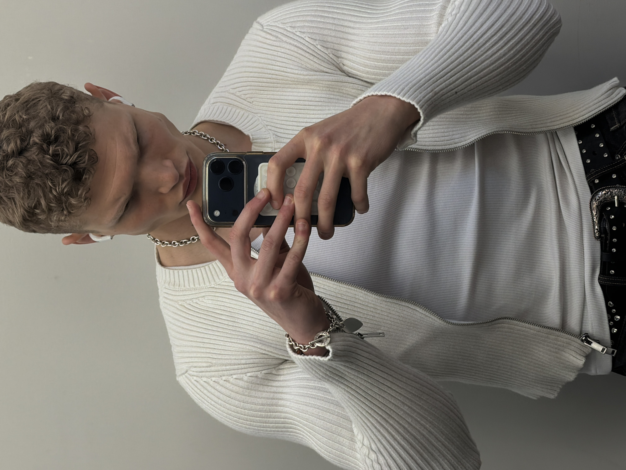 A young person, with short, curly, light brown hair, is depicted in a tilted, medium shot, appearing to take a mirror selfie or record themselves with a smartphone. They wear a white, ribbed, long-sleeved zipped cardigan, partially unzipped to reveal a plain white garment underneath. A thick silver chain necklace adorns their neck, and a silver bracelet with a heart charm and toggle clasp is on their left wrist. White in-ear headphones are visible in both ears.
The person holds a dark-colored smartphone, possibly an iPhone, with a clear case that has a white, circular accessory on the back. Their gaze is directed downwards towards the phone. On their right hip, a black studded belt with a prominent silver buckle is visible. The background is a plain, light-colored wall, suggesting an indoor setting. No specific time of day or external weather is discernible. The provided context for the location is London, United Kingdom, though no visual cues confirm this within the image. No clear text is visible on any of the subjects or objects.