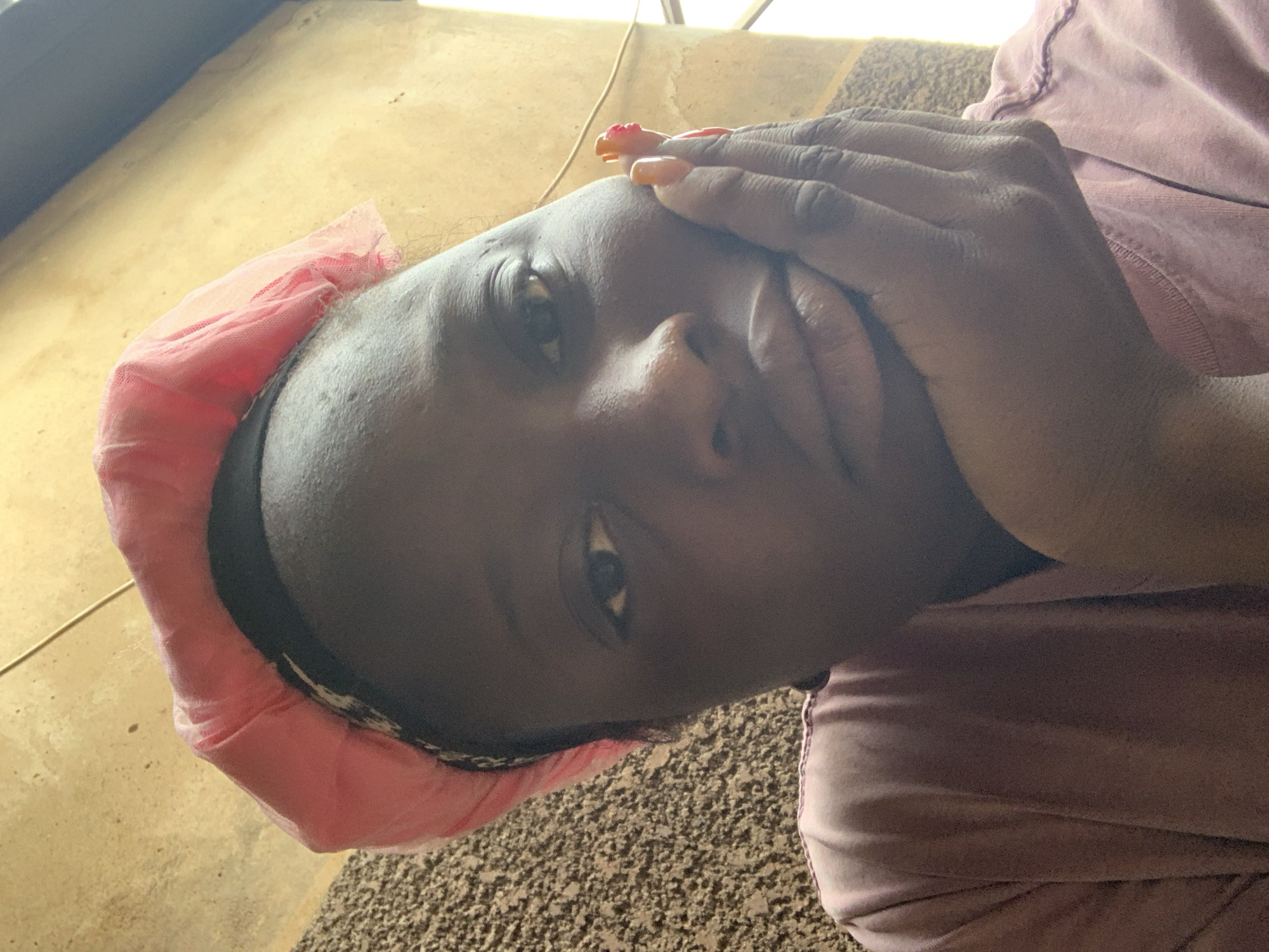 A close-up, slightly high-angle selfie features a dark-skinned person wearing a pink bonnet and a pink shirt. The person's left hand is supporting their chin, and their fingers are adorned with orange and pink nail polish. The background consists of a textured wall in shades of brown and beige, with a yellow wall and a light source visible on the right side. The image appears to have been taken indoors.