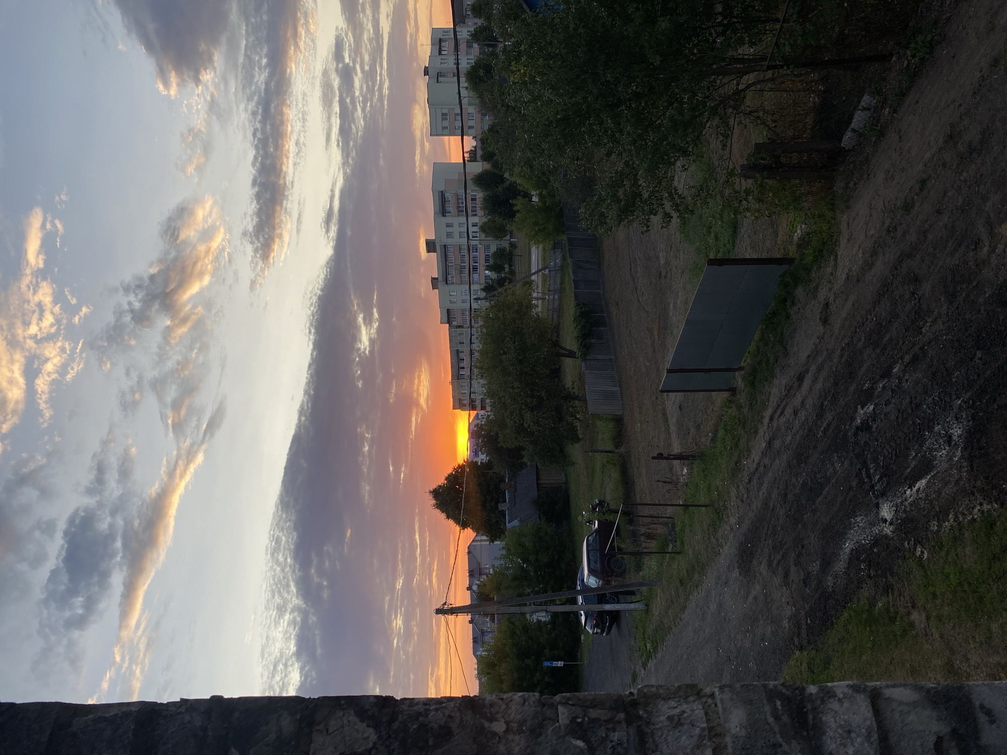 The image depicts a sunset scene in Krupki, Belarus. The sky is filled with dramatic clouds, tinged with orange and pink hues from the setting sun, which is visible on the horizon. In the foreground, a rough dirt path leads away from the viewer, with patches of grass on either side. A stone wall is visible on the far left, framing the scene.
In the middle ground, there is a collection of buildings and trees. Two cars are parked on a gravel area to the left, with a telephone pole nearby. Further back, a small wooden shed with a dark roof is nestled amongst trees. Beyond these, a row of apartment buildings stands tall, characteristic of Soviet-era architecture. A wooden fence runs across the middle of the scene, and further back, a metal fence and more trees are visible. The overall impression is of a rural or semi-urban environment at dusk. There are no people visible in the image.