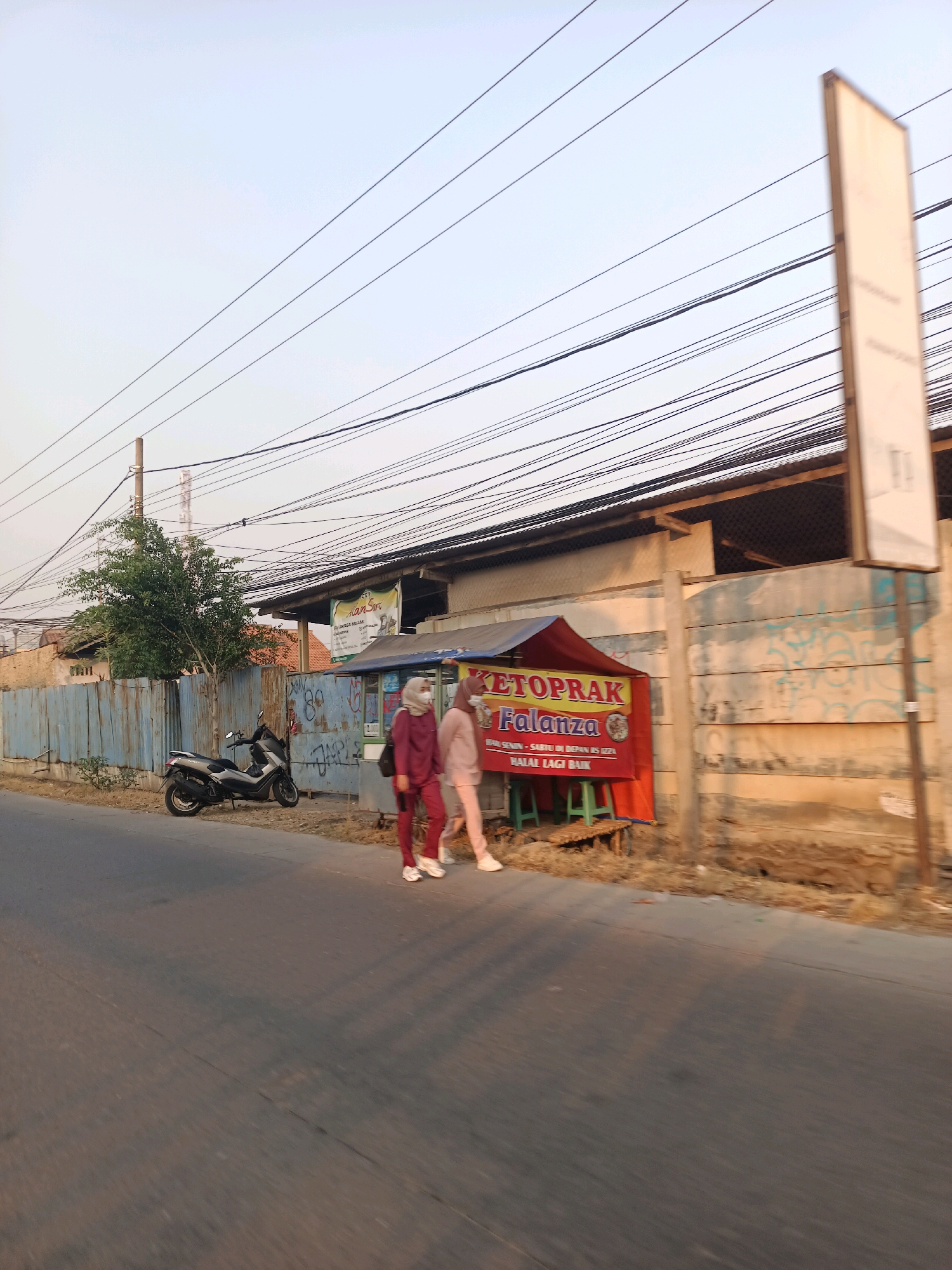 Two veiled women walk side-by-side on a sidewalk next to a street in Karawang, Indonesia. The woman on the left wears a burgundy hijab and matching outfit, while the woman on the right wears a pink hijab and matching outfit. They are walking away from the camera. Behind them is a small food stall with a red awning and a banner that reads "KETOPRAK Falanza". The stall has two green stools and a bamboo mat. Further back, there is a motorcycle parked next to a blue metal fence. A tree with green foliage stands behind the fence. The background shows a building with a corrugated metal roof and several utility poles with numerous wires running across the sky. The street is paved and appears to be a local road. The lighting suggests it is late afternoon or early morning.