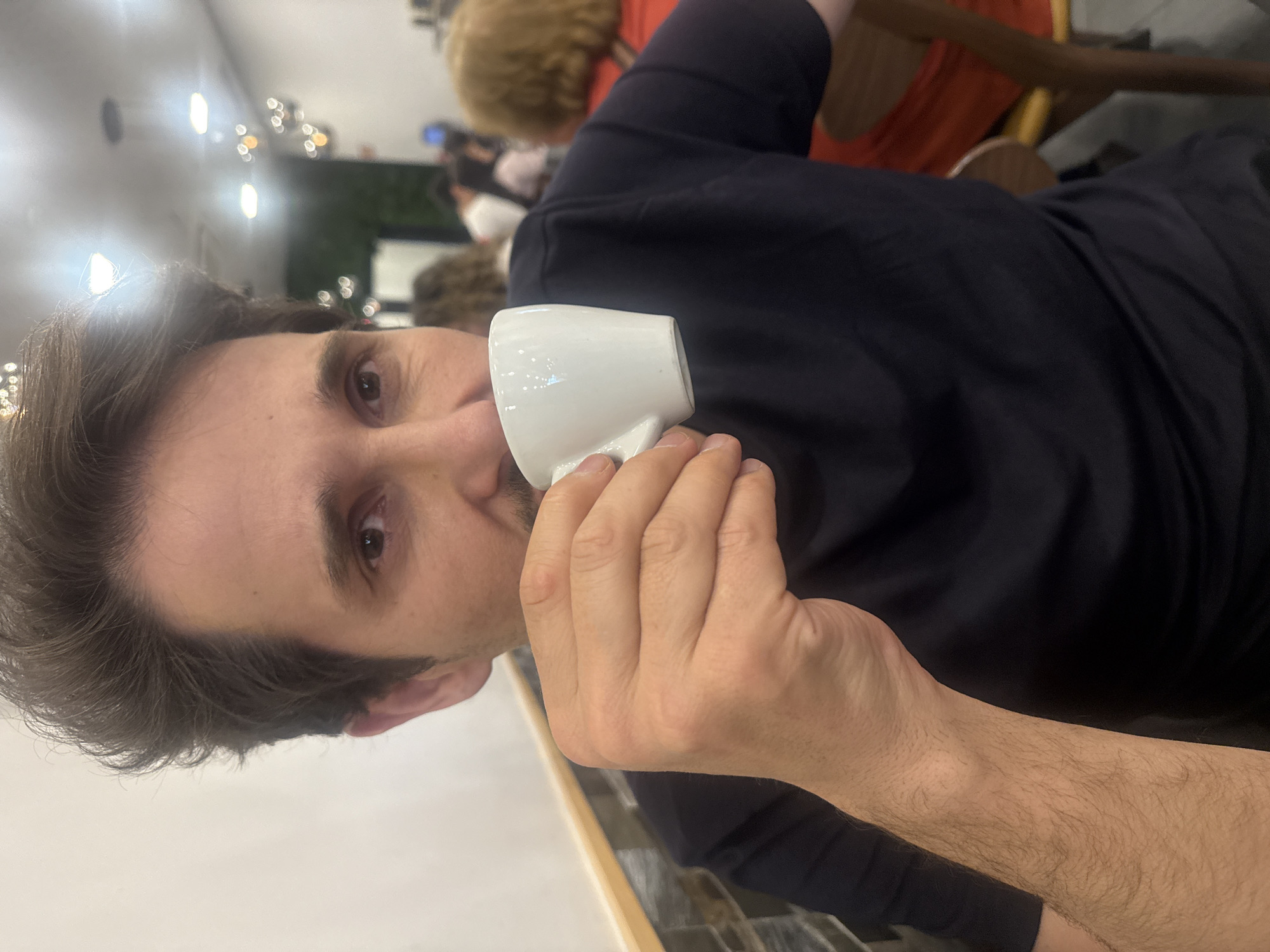 A man with short, dark hair and olive skin is shown drinking from a small, white cup. He is wearing a dark, possibly navy blue, t-shirt. He holds the cup with his right hand. In the background, there are other people and indistinct objects, suggesting an indoor public space. The lighting is somewhat bright and diffuse. The location is identified as Odivelas, Portugal, based on undisclosed information.