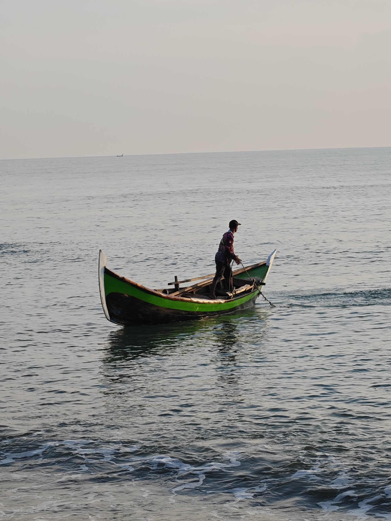 A lone fisherman stands in a small green and black wooden boat, which is partially submerged in calm, grey water. The man, wearing a dark baseball cap and a multi-colored shirt, is holding a rope attached to the boat and appears to be rowing or steering it. The boat is positioned in the middle of a body of water, with gentle ripples and a faint reflection of the boat on the surface. In the distance, a faint horizon line separates the water from a pale, overcast sky. The foreground shows smaller waves with white foam. This scene is likely taking place in the waters off the coast of Lhokseumawe, Indonesia.