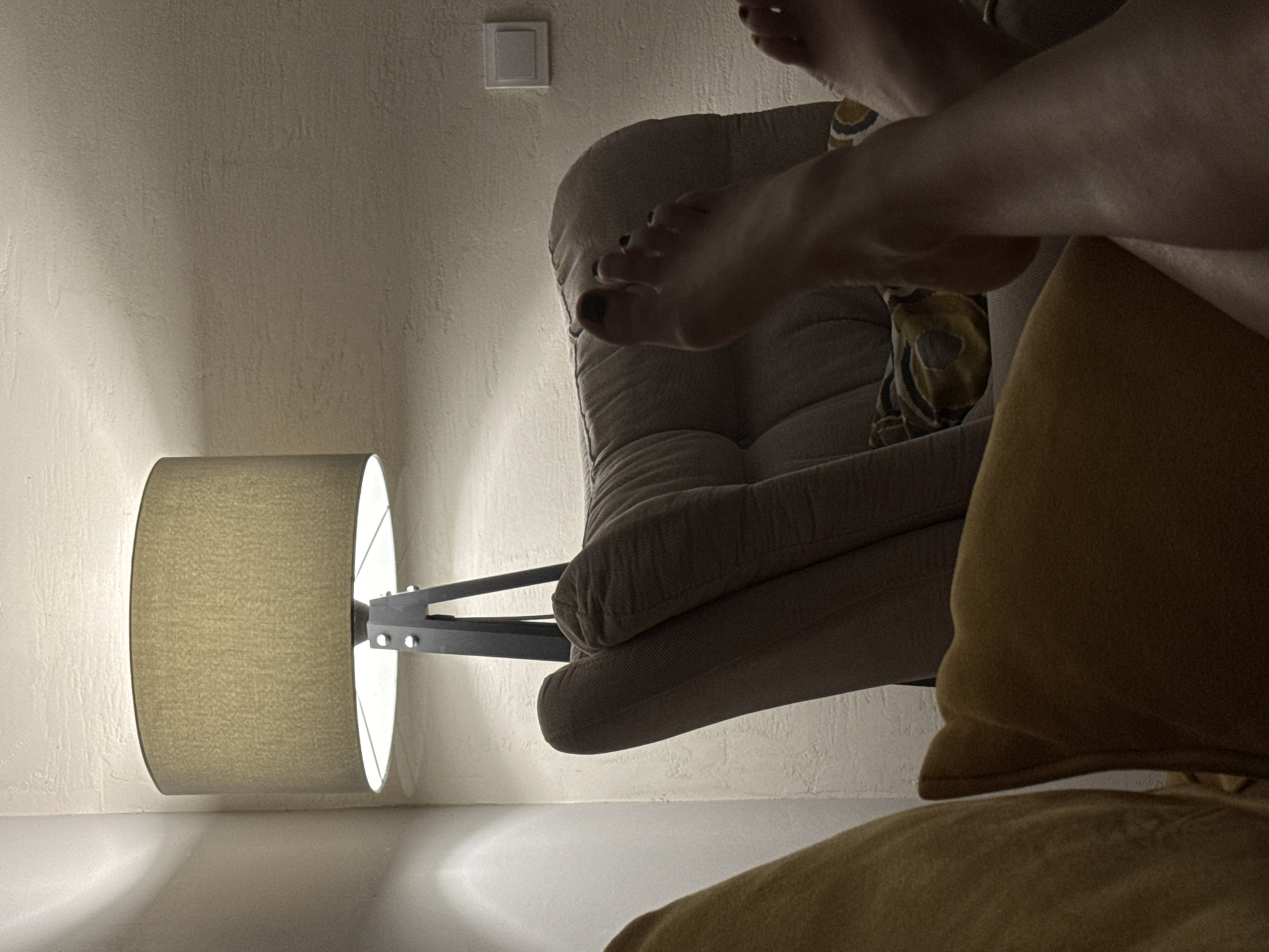 A dimly lit interior scene features a beige, cushioned armchair. A floor lamp with a beige lampshade stands to the left, casting light on the wall. A person's bare foot is prominently displayed in the foreground. A person wearing a yellow garment is seated in the armchair. A light switch is visible on the wall. The location is Podgorica, Montenegro.