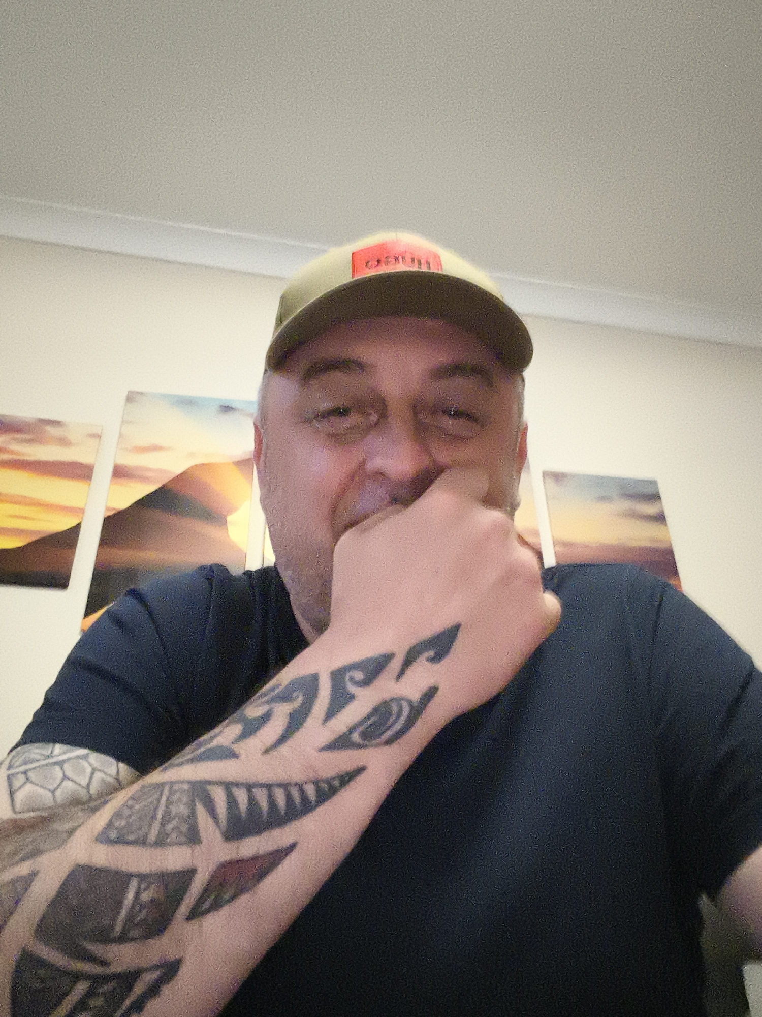 A middle-aged man is captured in a close-up, indoor portrait. He wears a dark, possibly navy blue or black, short-sleeved t-shirt and an olive green or khaki baseball cap with some red text on the front, partially visible as "ARTT" (likely "CARHARTT"). His right hand is raised, covering his mouth and lower nose, with knuckles visible, suggesting he is smiling or chuckling. His eyes are partially closed and crinkled at the corners, reinforcing an expression of amusement or happiness.
The man has extensive dark tattoos covering his right forearm and upper arm, featuring tribal or geometric patterns, including a prominent design resembling sharp teeth on his forearm. Another tattoo on his upper left arm, partially visible, has a hexagonal or scaled pattern. His hair is short and greyish-brown, and he has light stubble on his face.
The background consists of a plain, light-colored wall with three multi-panel canvas art pieces depicting a sunset or sunrise over mountains. The central panel is larger and shows a distinct mountain silhouette against a vibrant orange and yellow sky. A light-colored ceiling with a molding strip is visible at the top of the frame. The image appears to be taken indoors under artificial lighting. This individual is located in Huntingdonshire, United Kingdom.