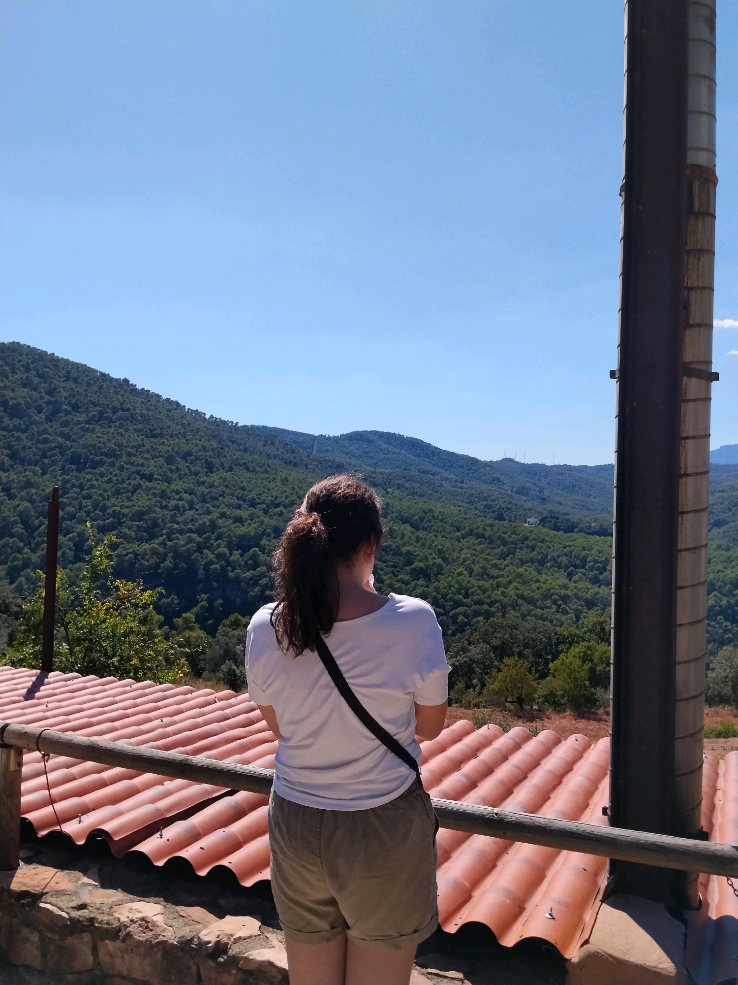 The image shows a person with their back to the camera, looking out at a vast, tree-covered mountain range under a clear blue sky. The person is wearing a white t-shirt and khaki shorts, with a black strap across their chest. In the foreground, there is a reddish-orange tiled roof and a wooden railing. To the right of the frame, a tall, dark cylindrical structure, possibly a chimney or a pipe, stands prominently. The overall scene suggests an outdoor viewpoint in a rural or mountainous area, likely in or near Caldes de Montbui, Spain, given the architecture of the roof tiles and the natural landscape.