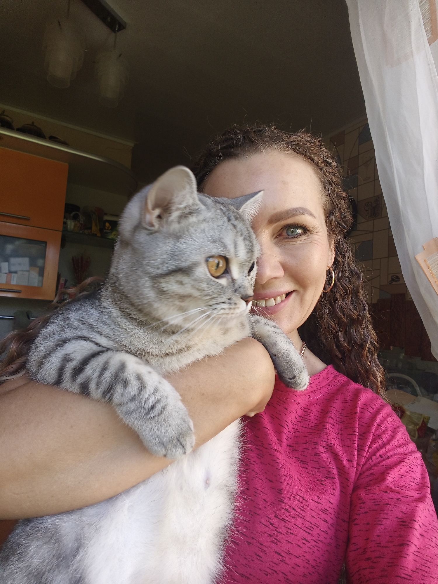 A woman with curly brown hair and green eyes is holding a grey tabby cat. She is smiling at the camera, and the cat is looking to the left. The woman is wearing a bright pink shirt. The background shows an indoor setting with some shelves and cabinets, suggesting a kitchen or living area. A white sheer curtain is visible on the right side of the image.