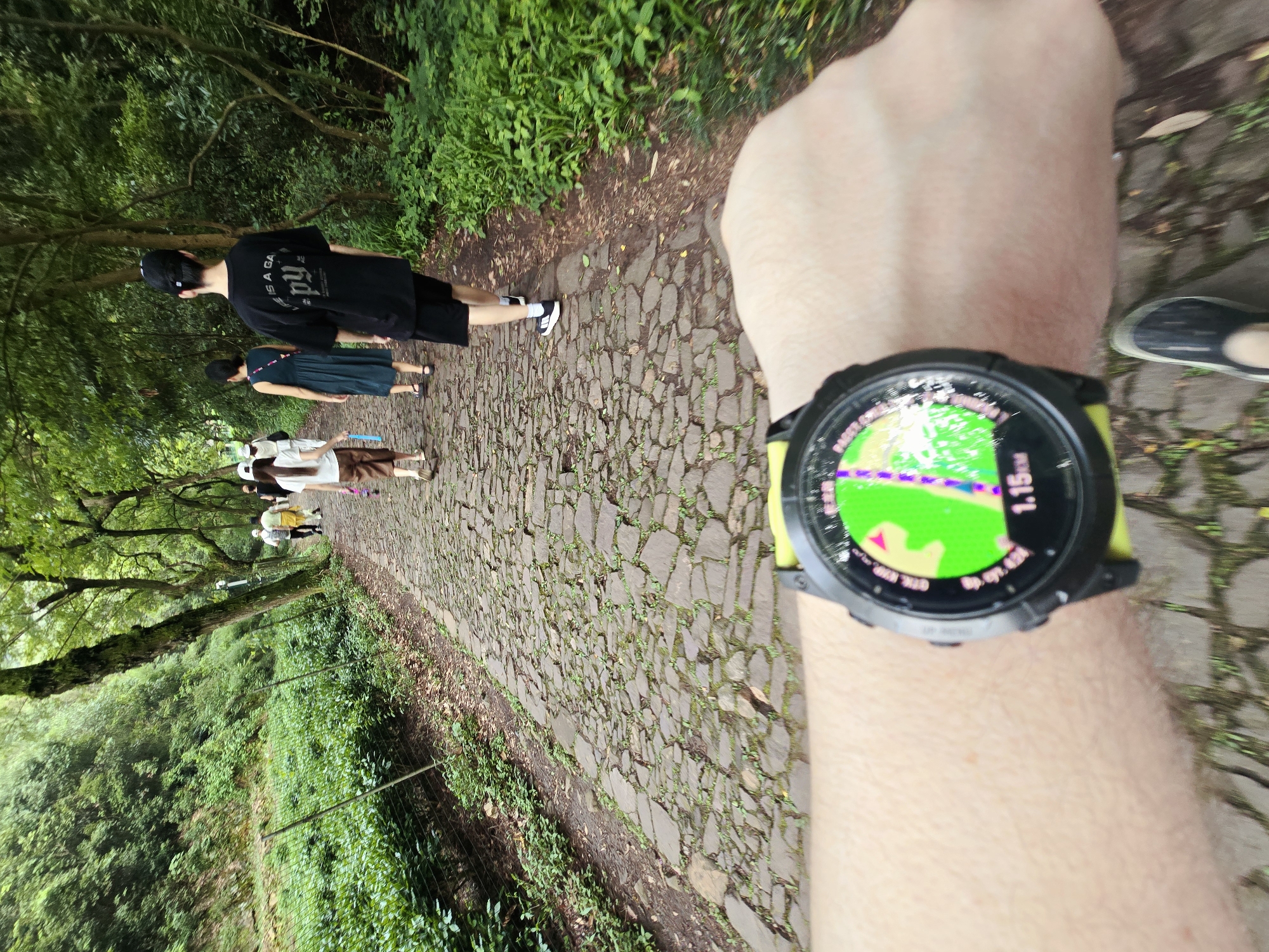 In Xihu District, China, a person's wrist wearing a smartwatch is prominently displayed in the foreground, showing a distance of 1.15 km and a map interface. The smartwatch has a yellow band. The person is wearing a dark shoe. The background depicts a stone-paved path winding through a lush green forest with trees lining both sides. Several people are walking along the path ahead. One person in the foreground wears a black t-shirt with text and black shorts, while another person in the middle ground wears a blue dress. Further ahead, individuals in lighter clothing are also visible. The environment suggests a natural, park-like setting, likely a hiking trail or scenic route within the district.