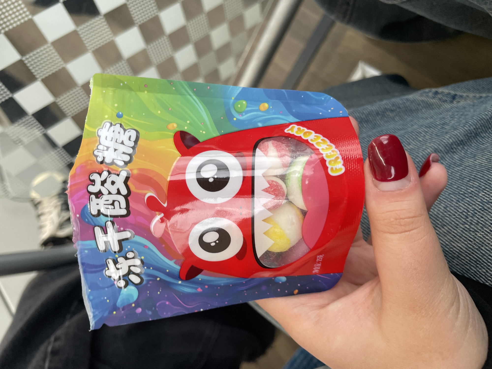 A person holds a colorful bag of candy with a cartoon monster design. The bag features Chinese characters and the text "FREEZEDRY". The candy inside appears to be a mix of round, colorful sweets. The background shows a checkered pattern and a person's lap dressed in denim. The image was taken in Yakovlevskoye, Russia.
