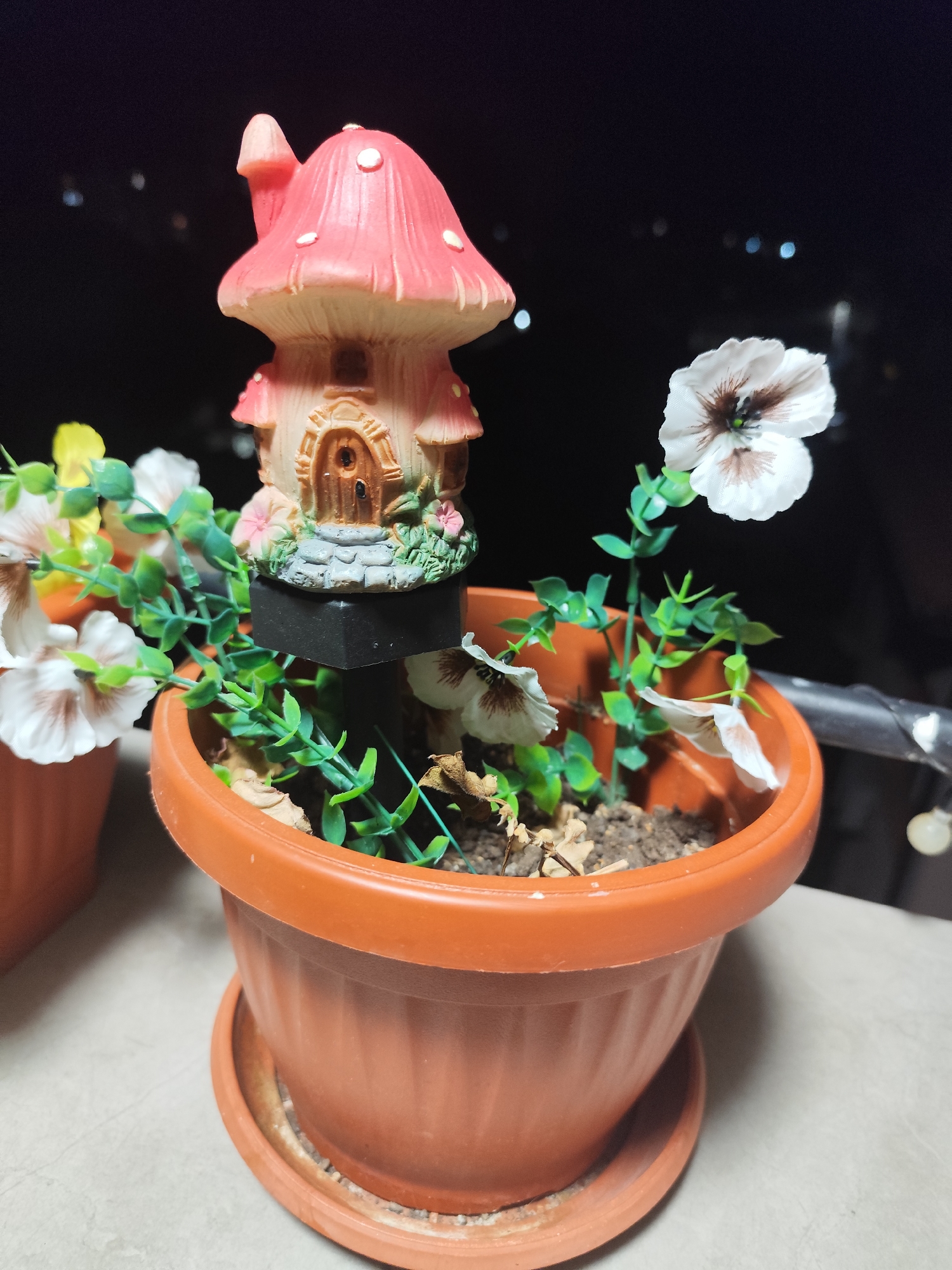 A close-up, eye-level shot shows a small, pink, mushroom-shaped decorative house, possibly a fairy house, placed in a brown plastic pot filled with soil and artificial greenery. The house has a pointed chimney, white dots on its cap, and a small arched wooden door with a tiny circular window. It sits on a black base. Several artificial white and yellow flowers with brown centers are scattered around the house. The pot is on a saucer and is placed on a light-colored surface, possibly a balcony railing, with a dark, out-of-focus background that suggests it is nighttime. The location is in Montana, Bulgaria.