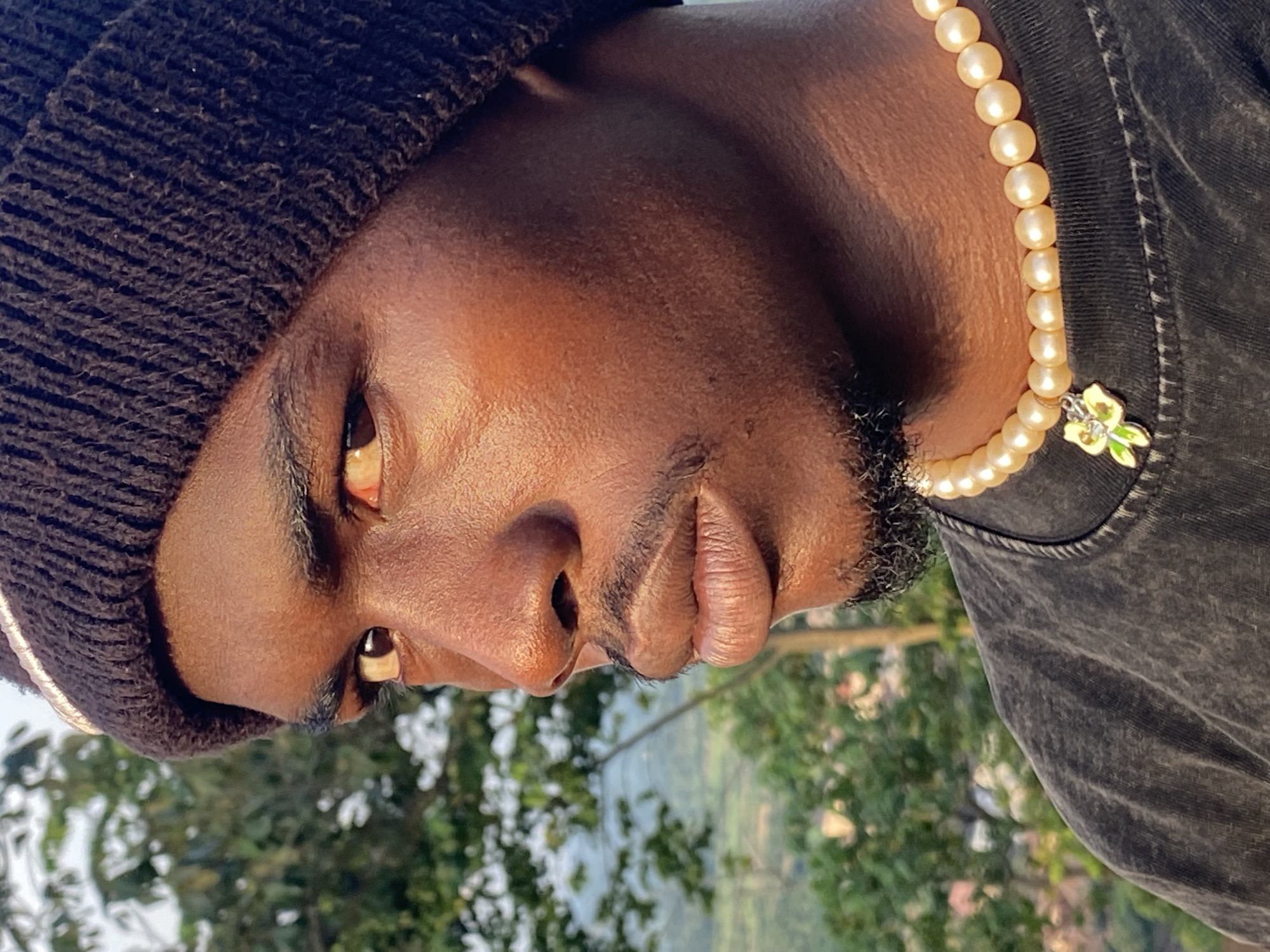 A close-up, profile shot shows a Black individual wearing a dark brown knit beanie and a grey, distressed t-shirt. The individual has dark eyebrows and a mustache, and a small amount of beard growth. Around their neck, they wear a pearl necklace with a small, green and yellow flower charm. The lighting suggests it is daytime, with the sun casting a warm glow on the individual's skin. In the background, out of focus, are green trees and a hint of a distant landscape, suggesting an outdoor setting likely in a non-urban environment, possibly on the outskirts of Abakaliki, Nigeria. The overall impression is a casual portrait.