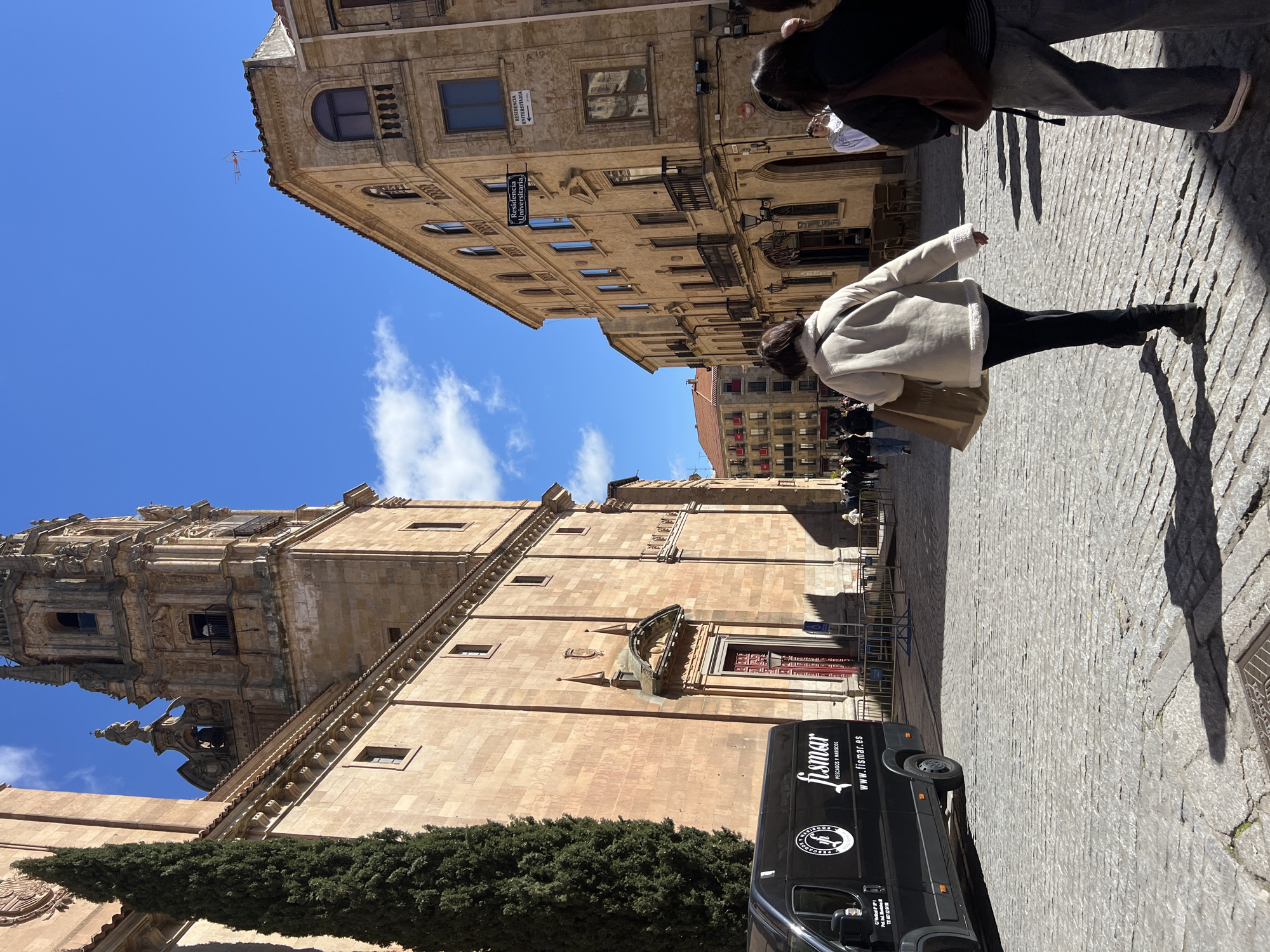 The image depicts a street scene in Salamanca, Spain, likely near the cathedral. The focus is on the architectural detail of the cathedral and the surrounding buildings, with a bright blue sky overhead. A person in a light-colored jacket and dark pants is visible walking away from the camera. A black van with the logo "fismar" is parked on the cobblestone street in front of the cathedral. The environment is sunny and suggests an urban setting within a historic district. Other pedestrians are visible in the background.