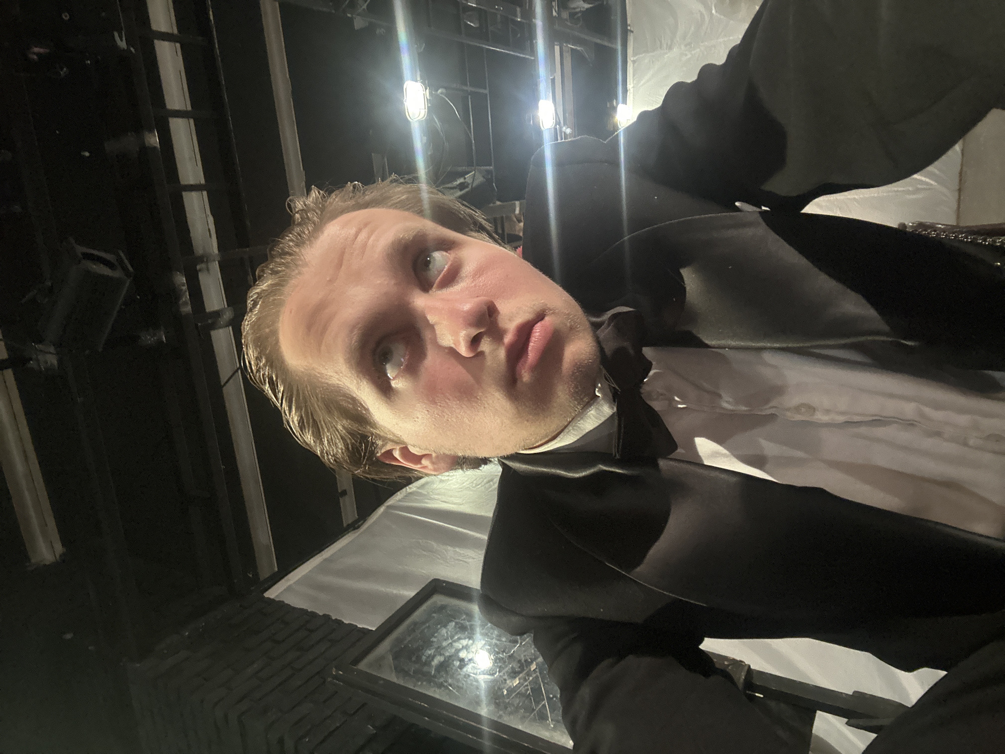 A young Caucasian male with blonde hair, wearing a black tuxedo jacket, a white dress shirt, and a black bow tie, looks upwards with a concerned expression. The setting appears to be backstage or a theater, with exposed beams, lighting fixtures, and white fabric drapes visible in the background. To the left, a dark, brick wall and a partial view of a window with a reflective surface are present. The lighting is dim, with bright spotlights casting shadows and creating lens flares. The overall mood is dramatic and somewhat unsettling.