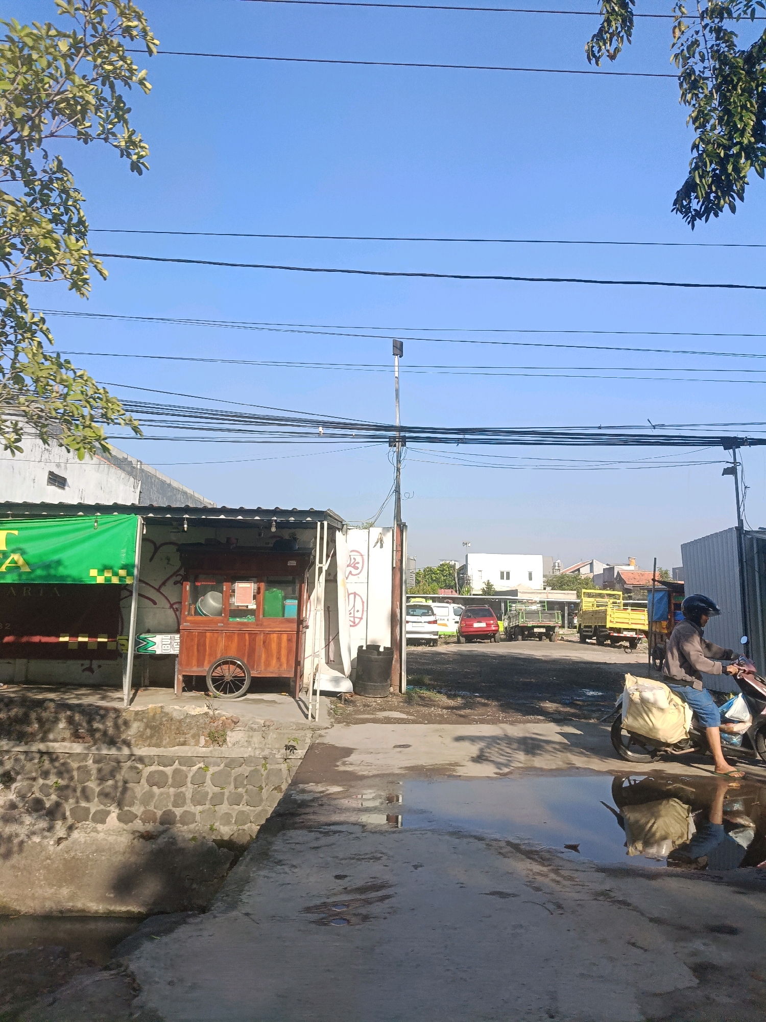 A sunny day scene depicts a street in Surabaya, Indonesia, with a clear blue sky overhead. Multiple power lines crisscross the sky. In the foreground, a concrete pathway with puddles of water reflects the surroundings. To the left, a food stall with a green awning and a wooden cart is visible. Behind the food stall, a wall with graffiti is present. Further down the street, several vehicles are parked, including cars and trucks. A person riding a motorcycle with bags strapped to the back is in motion on the right side of the image. The environment appears to be a mix of commercial and residential areas.