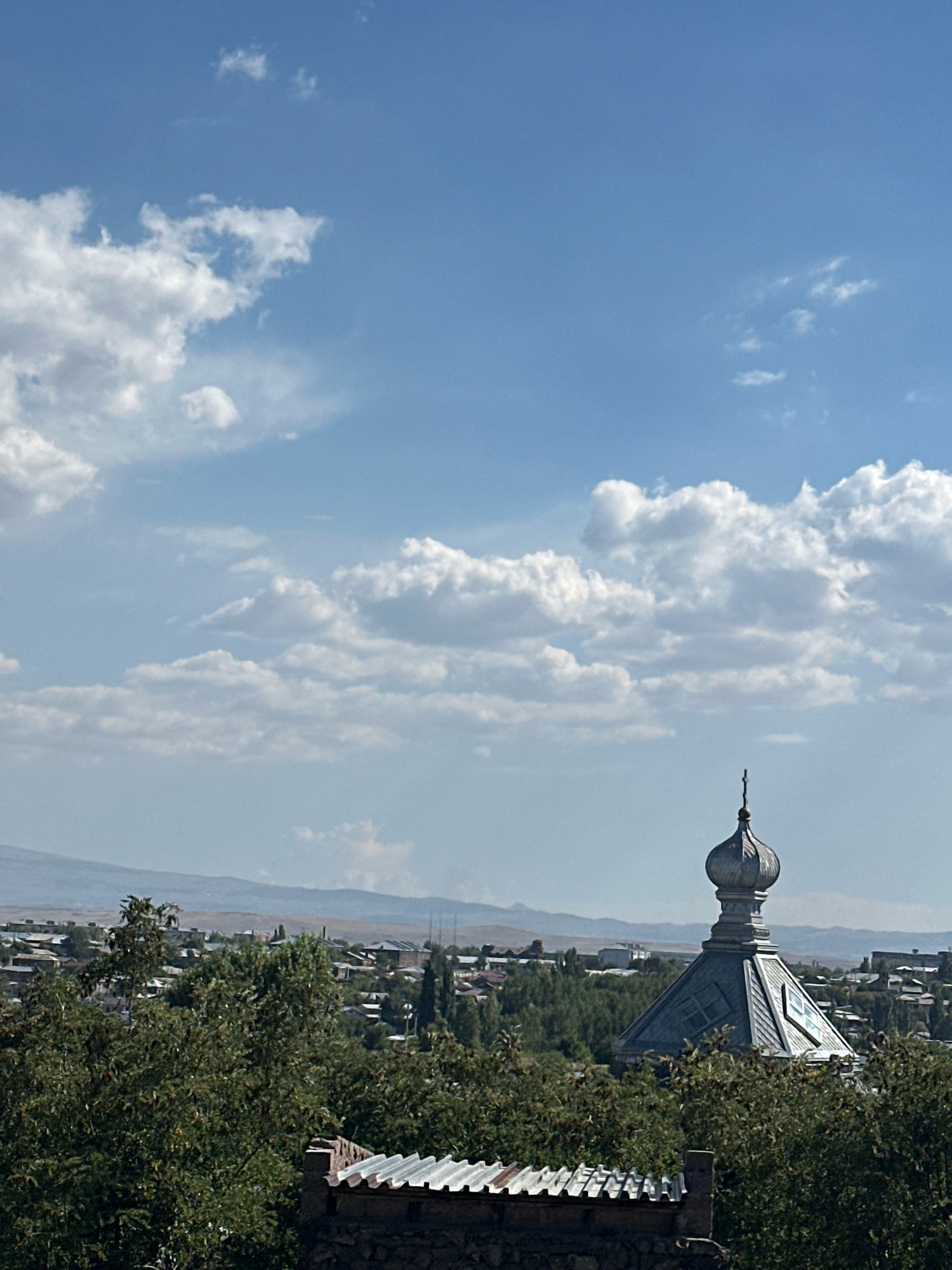 The image is a wide shot of a cityscape with a church dome in the foreground, taken from a slightly elevated position. The sky is a bright blue with scattered white clouds. The church dome, with a cross at its apex, is metallic and features geometric patterns and what appear to be skylights. Below the dome, the town stretches out, characterized by a mix of houses with red-tiled roofs and a dense canopy of green trees. In the distance, rolling hills and mountains are visible under a hazy sky. In the immediate foreground, a section of a stone wall and a corrugated metal roof are present, suggesting a viewpoint from a building or elevated structure. The scene is devoid of people or any visible activity, offering a tranquil view of the urban landscape of Gyumri, Armenia.