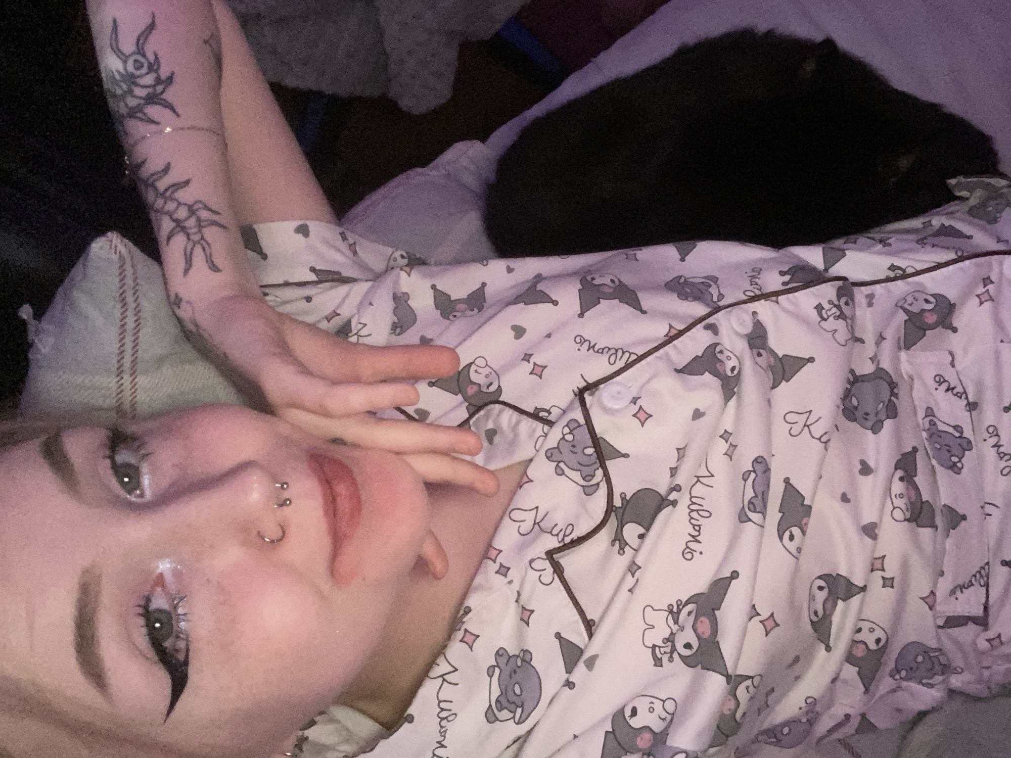 The scene depicts an indoor, dimly lit residential setting. No specific location within Izhevsk, Russia, is discernible. A single individual, appearing to be a young female, is shown in a close-up, upward-angled selfie. She is lying down, positioned against a light-colored surface, likely a bed or sofa.
The individual has light-colored hair, dark eyebrows, and wears visible makeup, including prominent black winged eyeliner, white glitter or shimmer in the inner corners of both eyes, and a light pink lip tint. Facial piercings include a silver septum hoop and two small silver studs in the left nostril. Her expression is a slight smile. Her left hand is partially visible near her chin.
She is wearing a white long-sleeved pajama top with a button-down front, featuring a repeating pattern of grey and black cartoon characters resembling Kuromi, interspersed with the text "Kuromi" and small pink diamond and heart symbols. The pajama top has brown piping along the placket.
Tattoos are visible on her left arm: a prominent black ink design resembling an elongated, multi-legged creature on the forearm, and a smaller, abstract symbol further up the arm. A delicate chain or bracelet is also present on her left wrist.
To the right of the individual, a dark-furred animal, consistent with a cat or small dog, is partially visible, resting on the same surface. A light-colored pillow with reddish-brown stripes is discernible behind the individual's head on the left side of the frame. The ambient lighting is low, with a faint pink or purple hue.