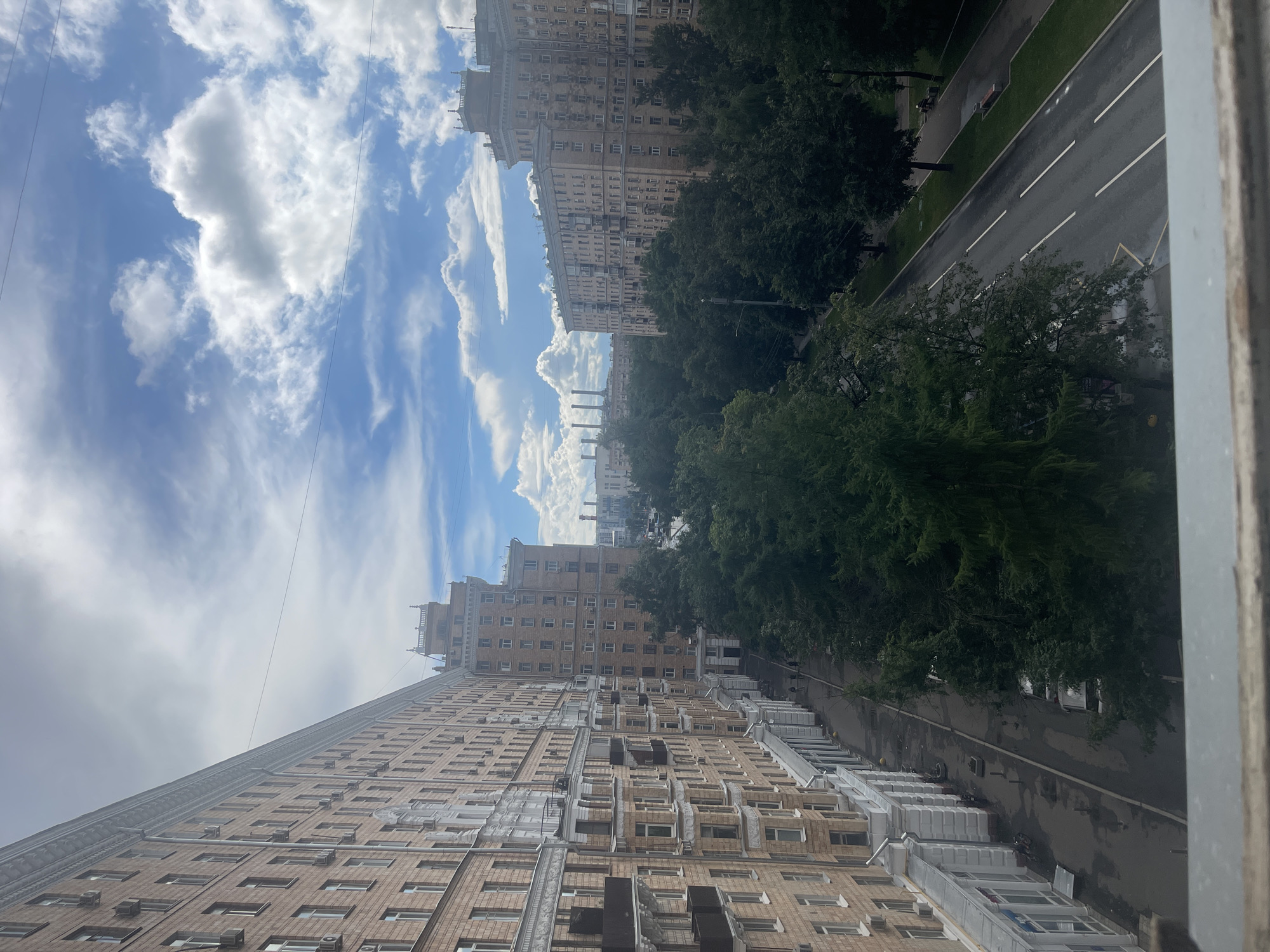 The image depicts a view of an urban street scene in Moscow, Russia, on a cloudy day. In the foreground, a tall, multi-story building with a light brown brick facade runs along the left side of the frame. The building features numerous windows and air conditioning units attached to its exterior. A sidewalk with awnings is visible at the base of this building.
Across the street, a tree-lined avenue is present, with dense green foliage dominating the midground. A two-lane road with white markings runs parallel to the trees. Further into the background, other large, similarly constructed apartment buildings can be seen, some with ornate architectural details at their upper levels. In the far distance, industrial structures with tall chimneys are visible, hinting at a power station or similar facility.
The sky is a mix of blue with dramatic white and gray clouds. There are power lines crisscrossing the sky. A few people can be seen walking on the sidewalk in front of the buildings. Parked cars are visible on the side of the road. The overall atmosphere suggests a residential area within a large city.