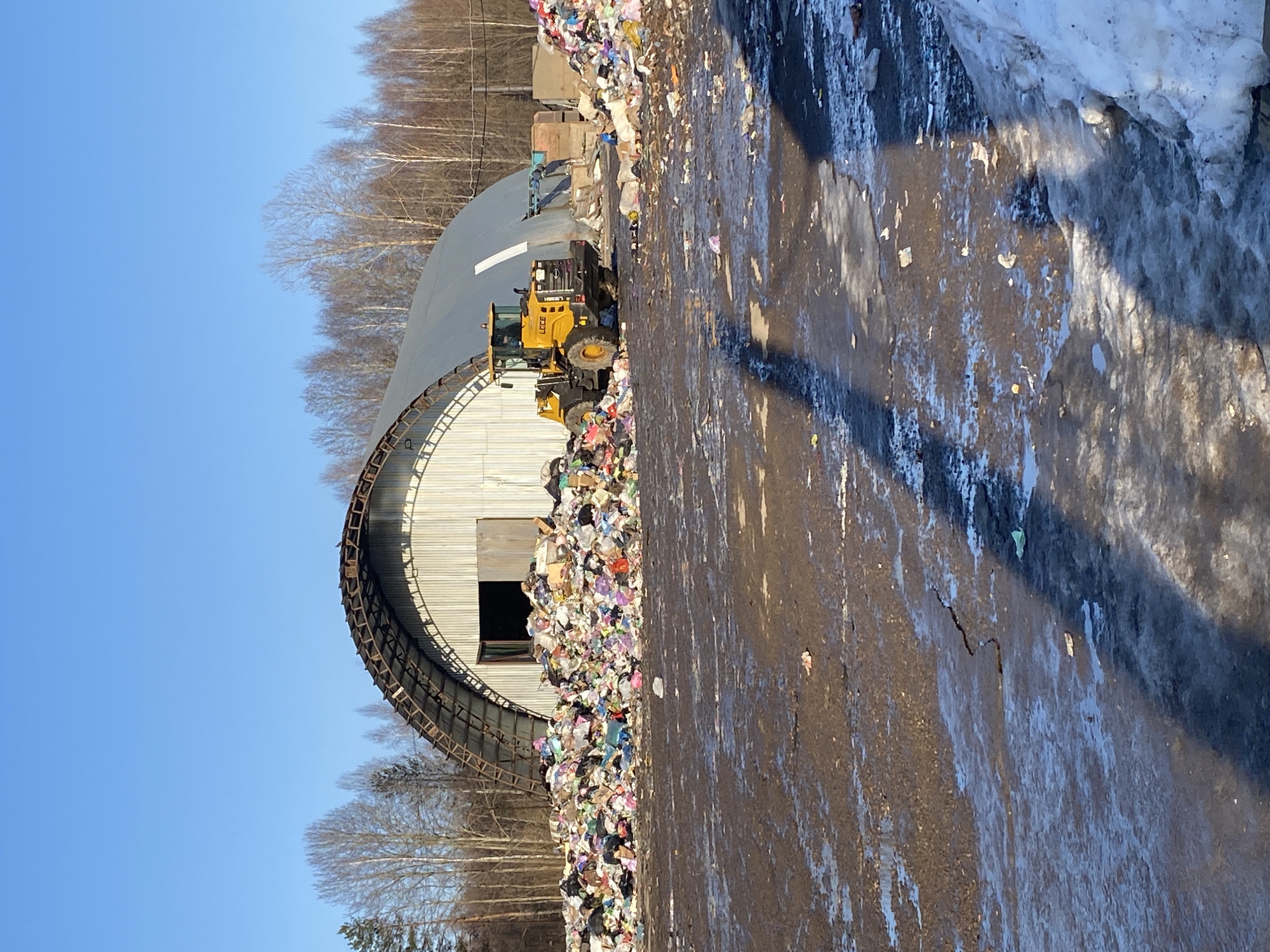 The image depicts a waste management facility near Ol'khovskoye, Russia. A large, arched metal structure stands next to a substantial pile of refuse, consisting primarily of plastic bags and assorted waste. A yellow front-end loader is positioned near the waste pile, likely used for moving and processing the refuse. The foreground shows a muddy or wet surface with scattered debris, and some patches of snow are visible on the right side, suggesting a cold climate. Bare trees are visible in the background, further indicating a winter or early spring setting. The sky is clear and blue.