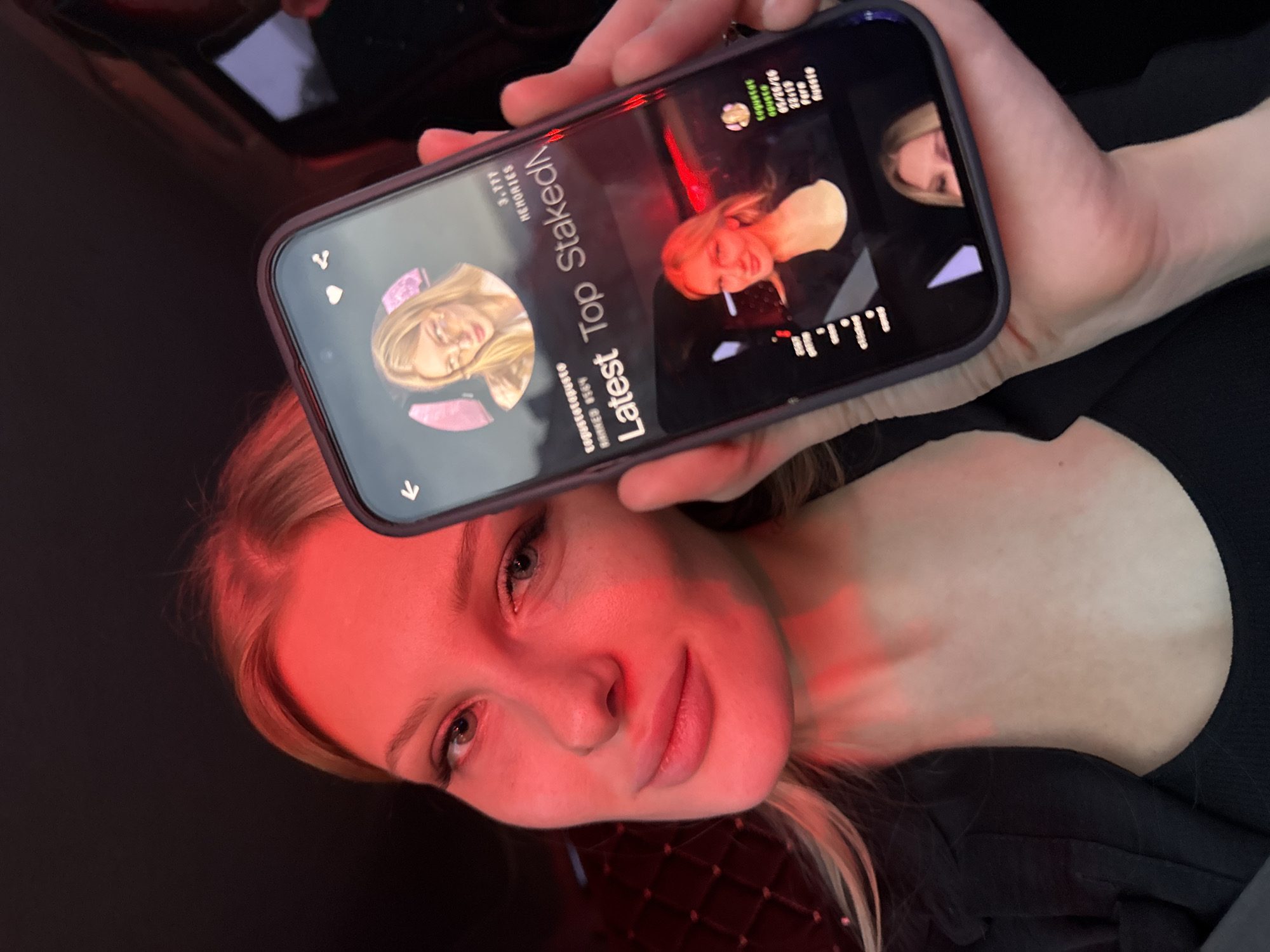 A fair-skinned blonde woman is holding a smartphone in front of her face. The screen displays a social media profile with the text "Latest Top Staked," her rank of 8564, and "Memories - 3.777." The woman's face is illuminated with a red light source. The background is dark, and the setting is likely indoors, possibly in a car. The location is unknown and cannot be inferred from the image.