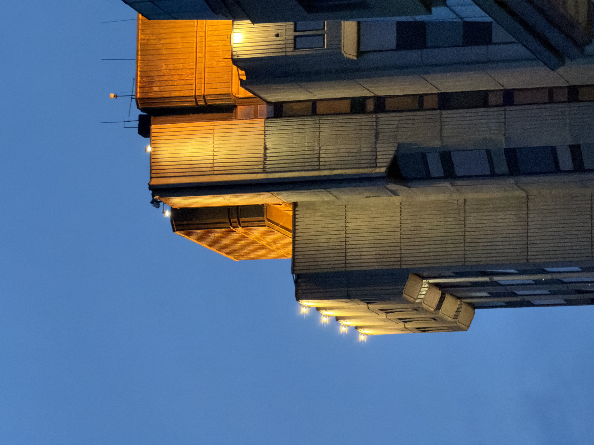 This image shows the architecture of a building against a blue sky. The building features a repeating pattern of gray horizontal panels. Some sections have orange accents and multiple small lights are visible. The photograph is taken from a low angle. There is no indication of the location being Varna, Bulgaria.