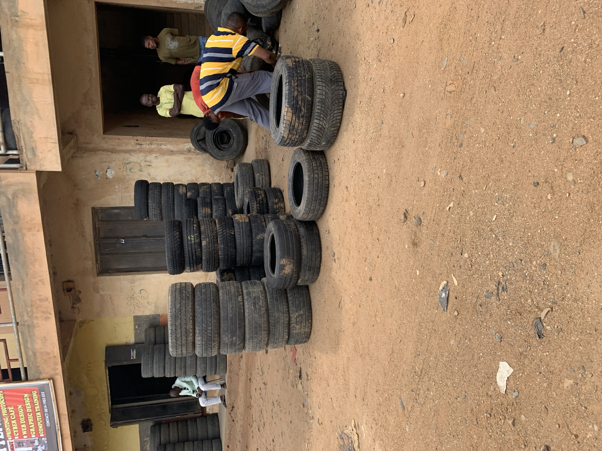 The image shows a dirt lot adjacent to a multi-story building. Stacks of used tires are present in the lot and inside the building, suggesting a tire shop or storage facility.
Several people are visible:
- One man is crouching near a stack of tires, examining them. He is wearing a horizontally striped yellow and blue shirt and gray pants.
- Two men are standing in a doorway inside the building. One is wearing a yellow shirt, and the other is wearing a green shirt.
- Another man is sitting on the ground, partially obscured by a stack of tires. He is wearing a light green shirt and white pants.
The building appears to be in a state of disrepair.
The location is Rafin Par, Nigeria.
