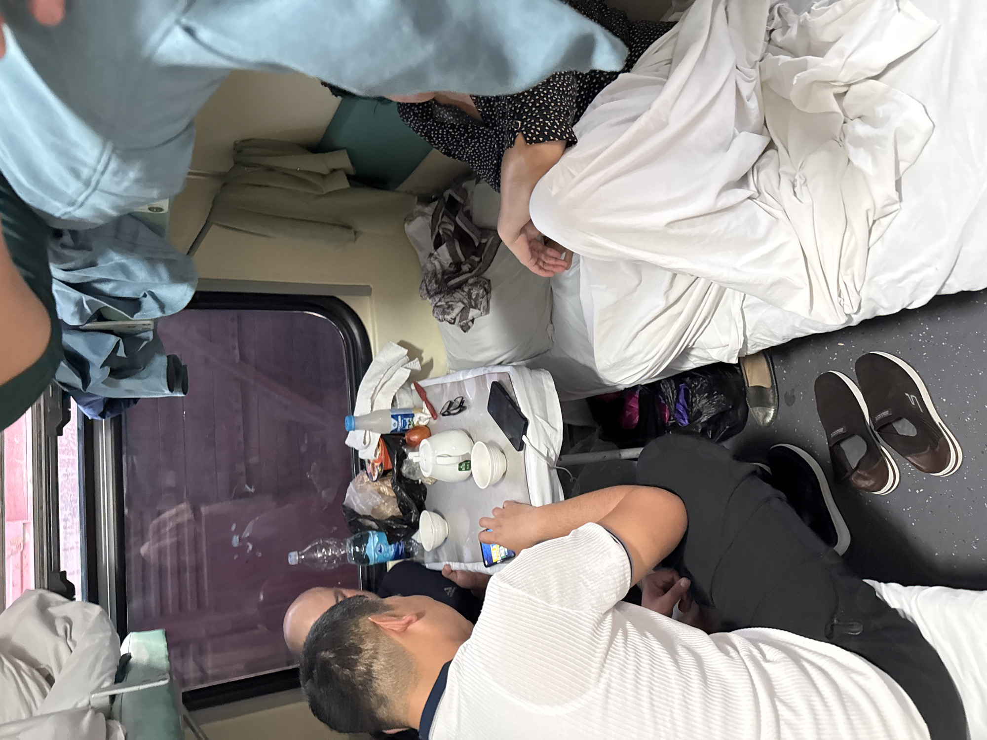 The image depicts the interior of a train cabin. Two people are in the lower berth, one lying down and looking at a phone. A second person is sitting and also looking at the phone. Various personal items are on a small table, including a tea set, water bottles, and a smartphone. The berth above appears to have clothes and blankets on it, and someone is sleeping on the upper berth. The scene suggests travel, and there is a window behind the seated person. The location is unknown.