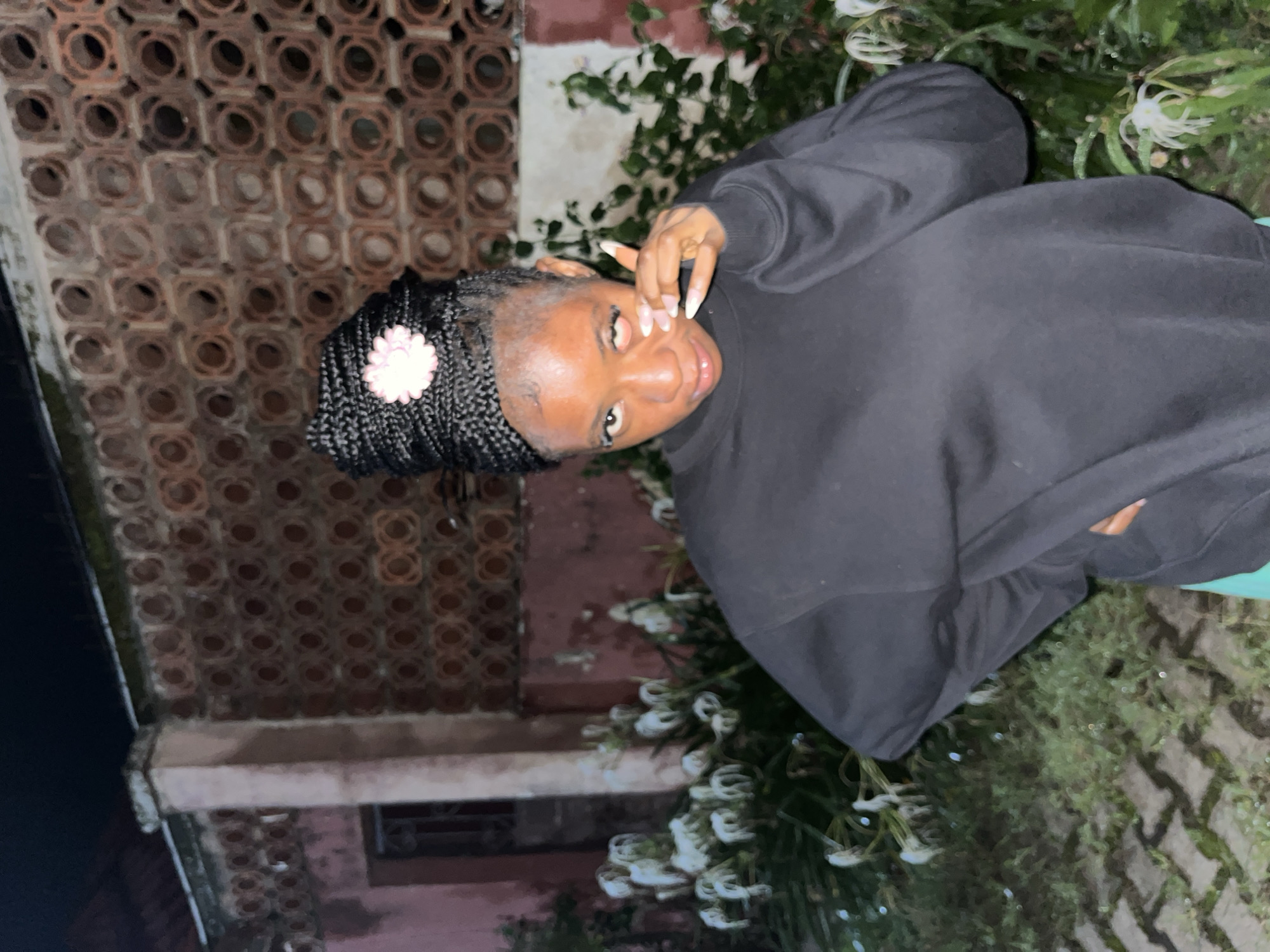 A young Black woman with braided hair, adorned with a pink flower accessory, is captured in a medium shot. She is wearing a black sweatshirt and green shorts. Her left hand is placed on her cheek, and her right hand is on her hip. The background features a wall constructed with circular patterned terracotta bricks, a pink-painted pillar, and lush green foliage with white flowers. The scene appears to be outdoors, possibly in a garden area, with a cobbled pathway visible in the foreground. The lighting suggests it is nighttime, and the overall ambiance is casual. The location is identified as Jos, Nigeria.