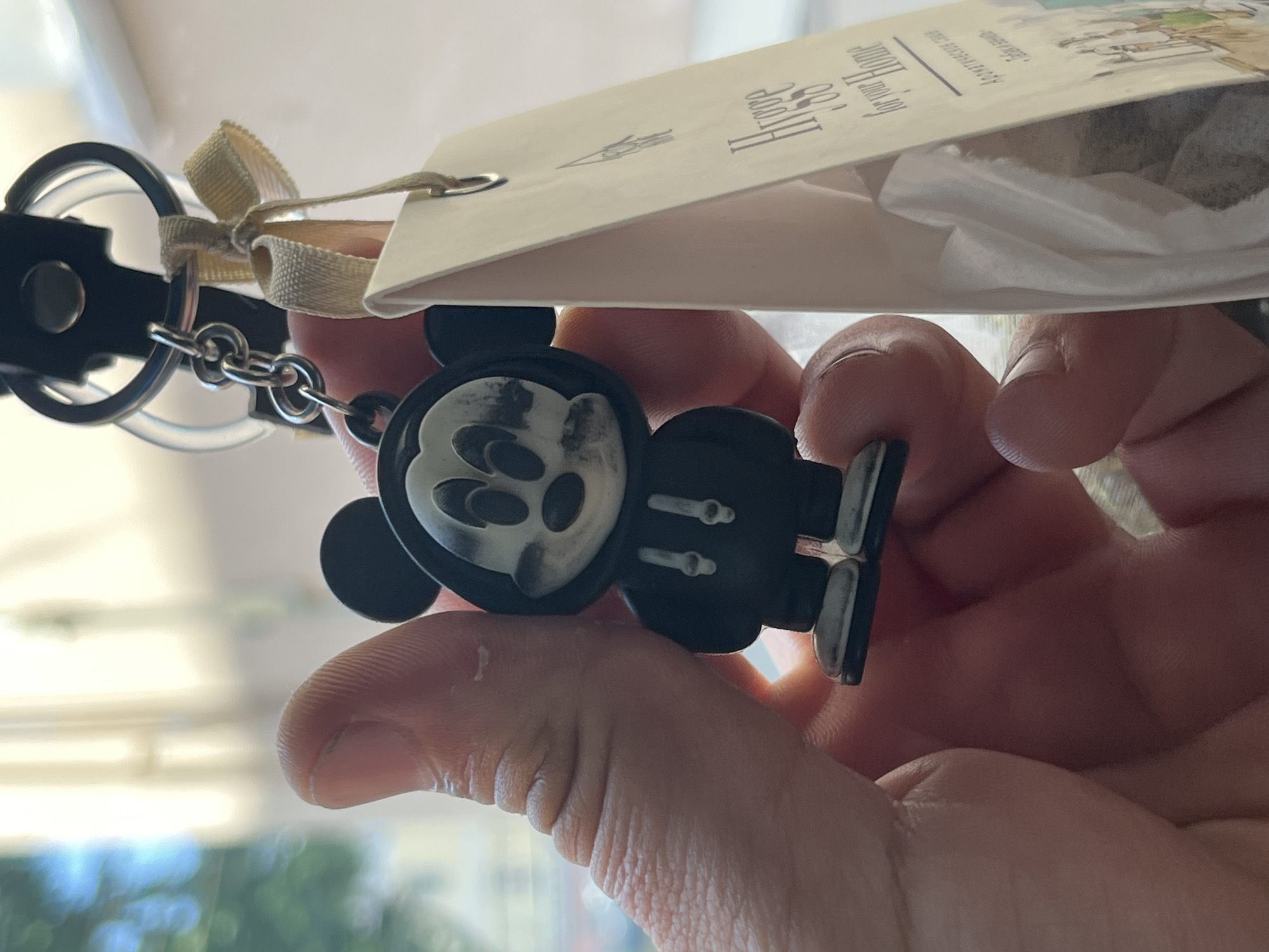 In Kazan, Russia, a person's hand is holding a Mickey Mouse-themed keychain with a black hoodie. A light-colored tag is attached to the keychain, with text that includes "Hygge for Your Home" and some Russian text. The background is blurred, showing hints of daylight and possibly buildings.