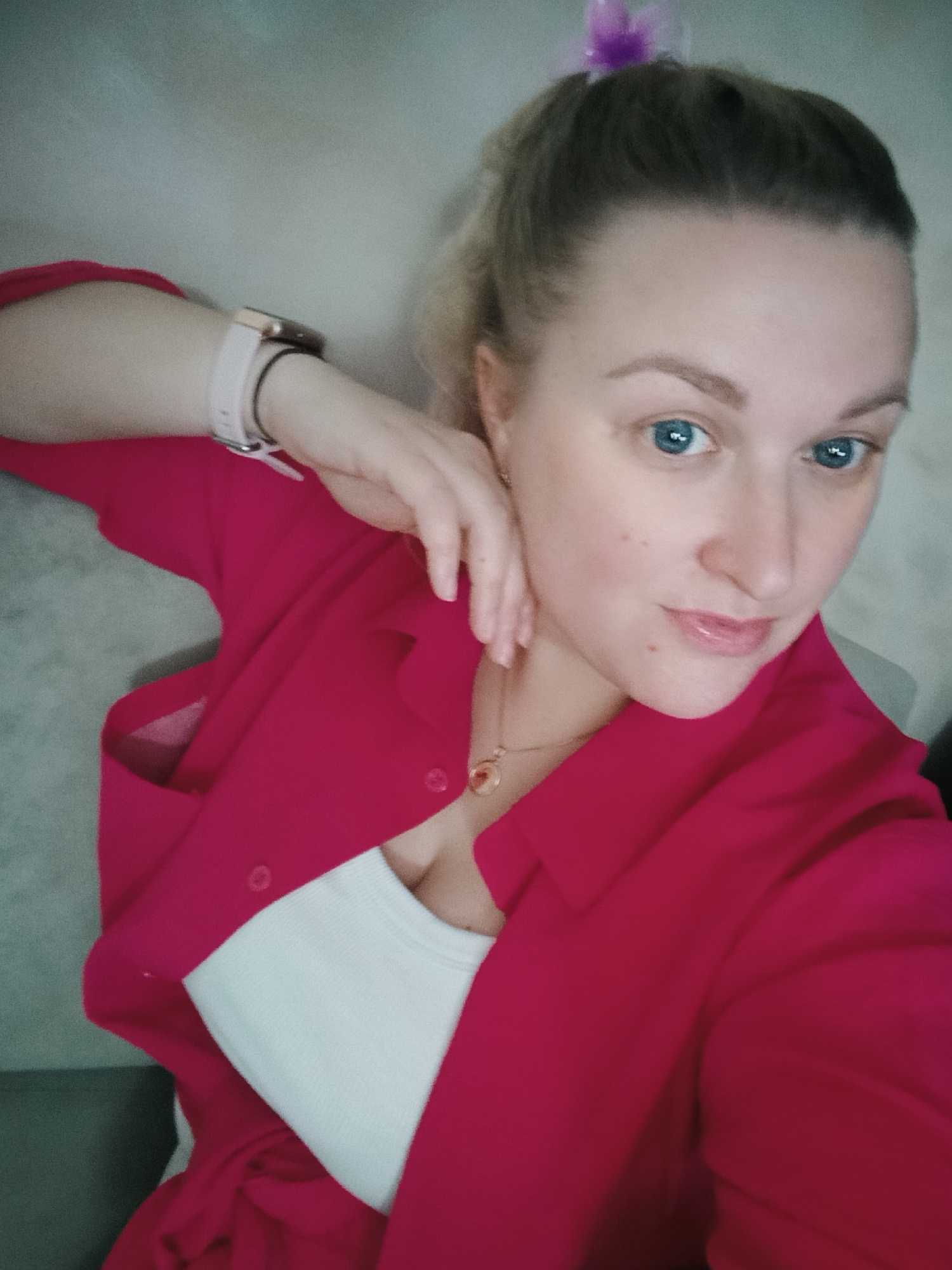 A fair-skinned woman is depicted in a close-up selfie, looking directly at the camera with striking blue eyes. Her hair, appearing light blonde or brown, is pulled back into a ponytail, secured with a small purple hair accessory visible at the top of her head. She wears natural-looking makeup, including defined eyebrows and light pink lips, with a few small moles or freckles on her cheek.
She is dressed in a vibrant magenta or hot pink long-sleeved shirt or light jacket, worn unbuttoned over a white ribbed tank top. On her left wrist, she wears a light-colored smartwatch with a metallic casing. A delicate gold-toned chain necklace with a small pendant is visible around her neck. Her right hand is gently raised, resting near her face and neck. The background is a simple, blurry, light-colored indoor surface. Her expression is calm and friendly, with a subtle, pleasant smile. The image was taken in Chaikovsky, Russia.