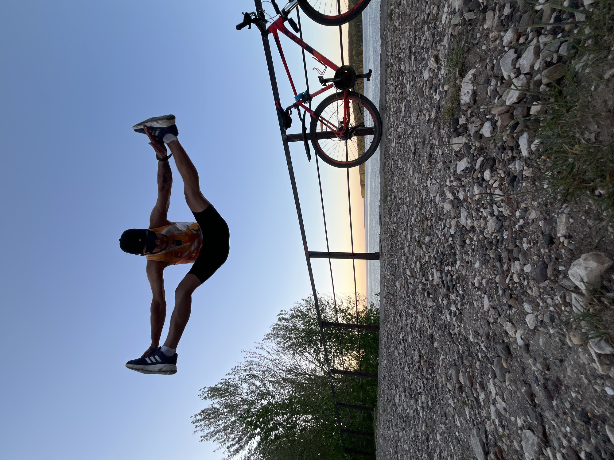 A man in a yellow and black athletic outfit with a black baseball cap is standing on his hands with his legs extended upward, reaching toward his head and his feet pointing to the sky. He is wearing blue athletic shoes. A red and black bicycle is leaning against a metal railing, which overlooks a body of water with a sunset. The railing is adjacent to a gravelly area. The background features trees and a sky transitioning from light blue to a gradient of sunset colors. The image is taken from a low angle, resulting in a rotated perspective. The location is Chaikovsky, Russia, based on visual recognition.