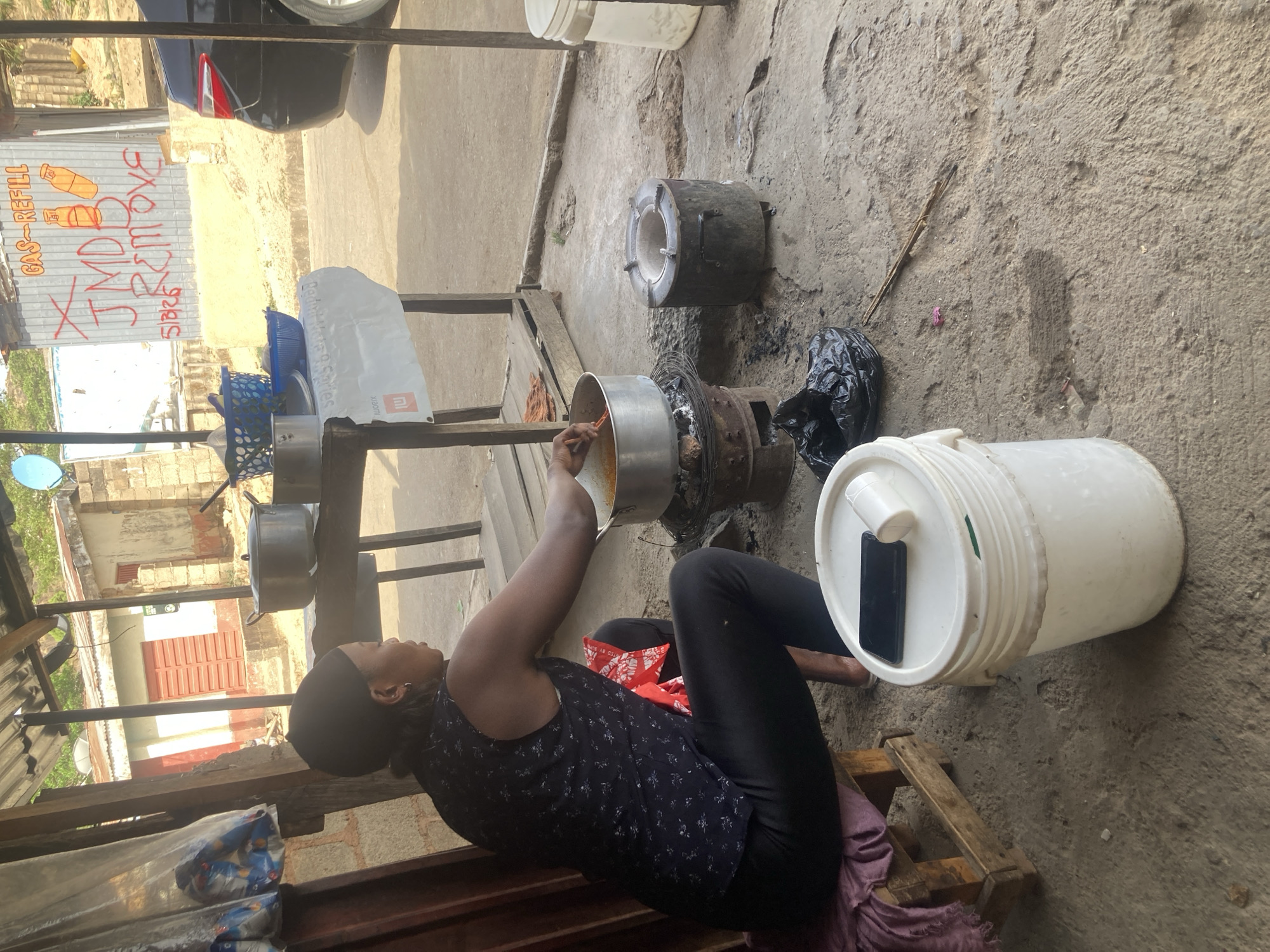 The image captures an outdoor scene in Jos, Nigeria, depicting a person engaged in cooking during the daytime. A person, with their head covered by a dark fabric, is seated on a low wooden pallet, stirring a large silver pot placed on a traditional charcoal or wood-burning stove situated on the rough concrete ground. An identical, empty stove sits nearby.
Around the cooking area, a white plastic bucket with a black smartphone resting on its lid is visible, alongside a black plastic bag on the ground. In the background, a corrugated metal fence or wall displays red graffiti. The text "GAS-REFILL" is prominently visible, accompanied by drawings of two gas cylinders. Below this, partially legible text includes "XMDB REMOVE" and what appears to be "5196".
Further in the background, simple concrete structures with red window shutters and a small satellite dish can be seen. The rear portion of a black car is visible at the top of the frame. Bright sunlight indicates it is daytime, casting clear shadows. A white paper bag with a "xiaomi" logo is also present on a makeshift shelf. The scene suggests an informal cooking setup, possibly a small business or household activity.