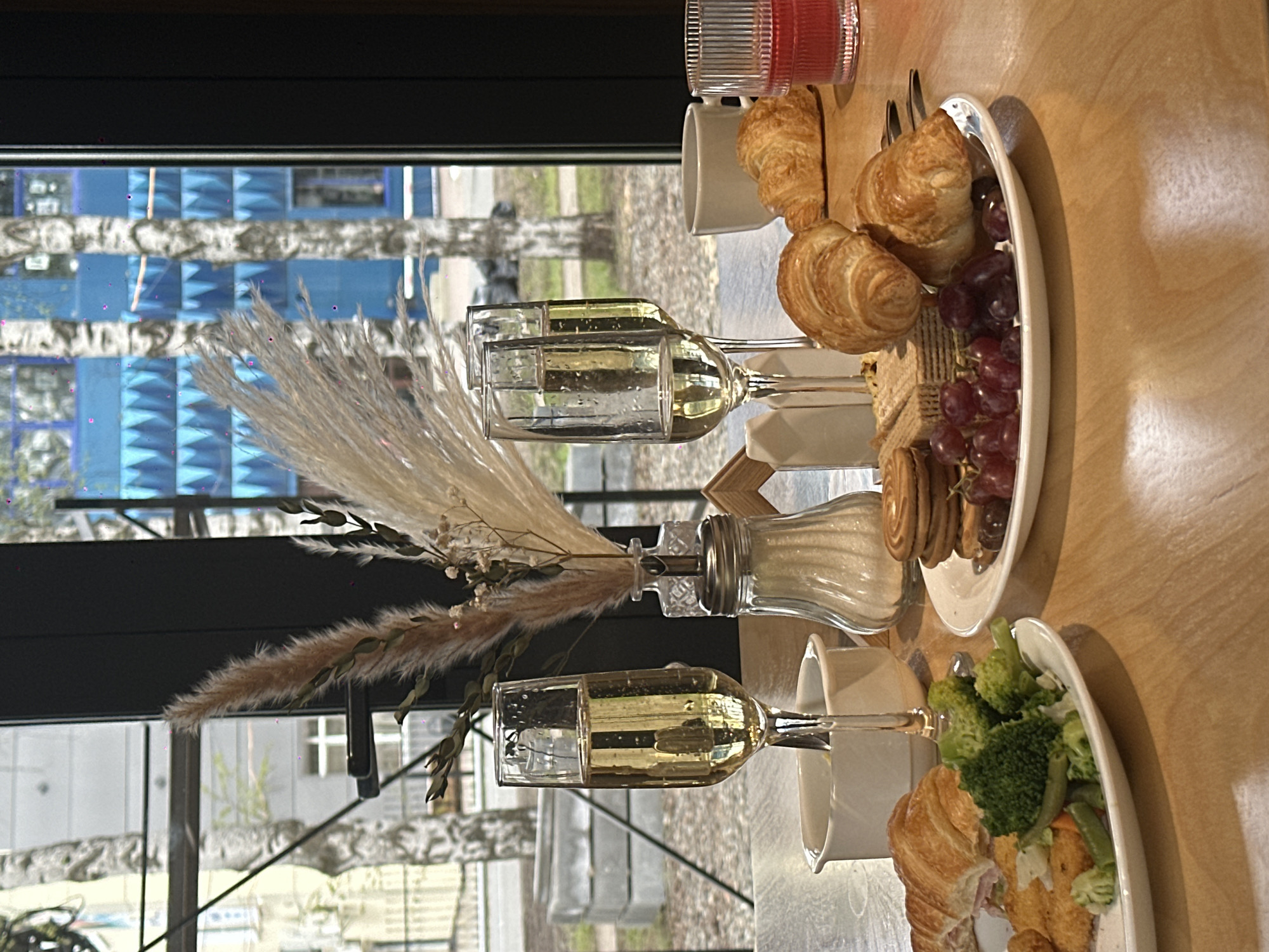 The image captures a breakfast scene set on a wooden table, likely in an establishment. Two champagne glasses filled with golden liquid stand alongside cups. One plate contains croissants, crackers, cookies, and grapes. Another plate features a croissant sandwich and a mix of vegetables, including broccoli. In the background, a window frames an urban view, hinting at the possible location of Glazov, Russia, based on architectural styles and the prominence of birch trees, although precise geolocation cannot be determined from the visual information alone. There are no visible people or explicit interactions in the image, focusing instead on the still life of a morning meal setting.